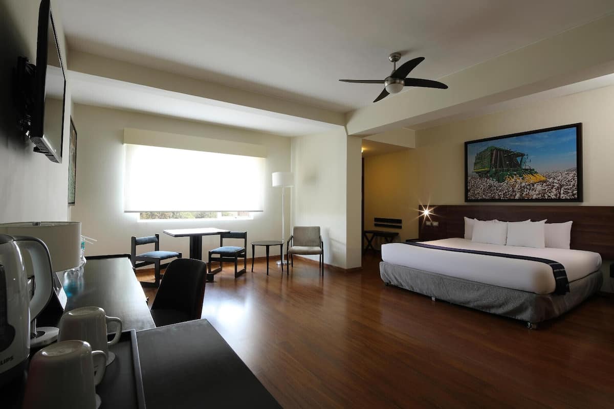 A hotel room with a bed, desk, chairs, a coffee machine, and a painting on the wall.