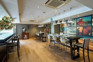Interior de una cafetería con suelo de madera, asientos en la barra, iluminación colgante y una pared decorada con obras de arte de colores.