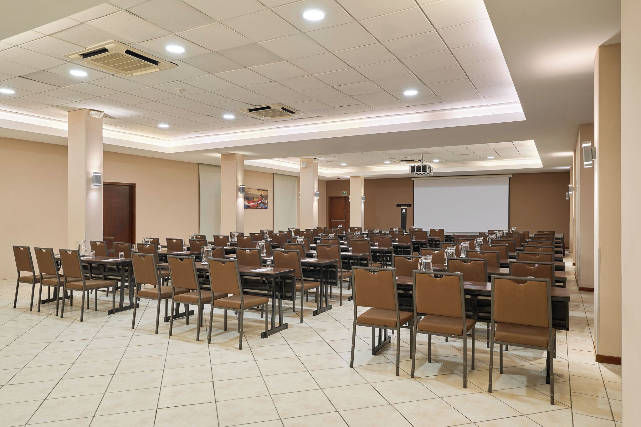 Un salón de conferencias con varias mesas y sillas, una pantalla de proyector y una gran pizarra.