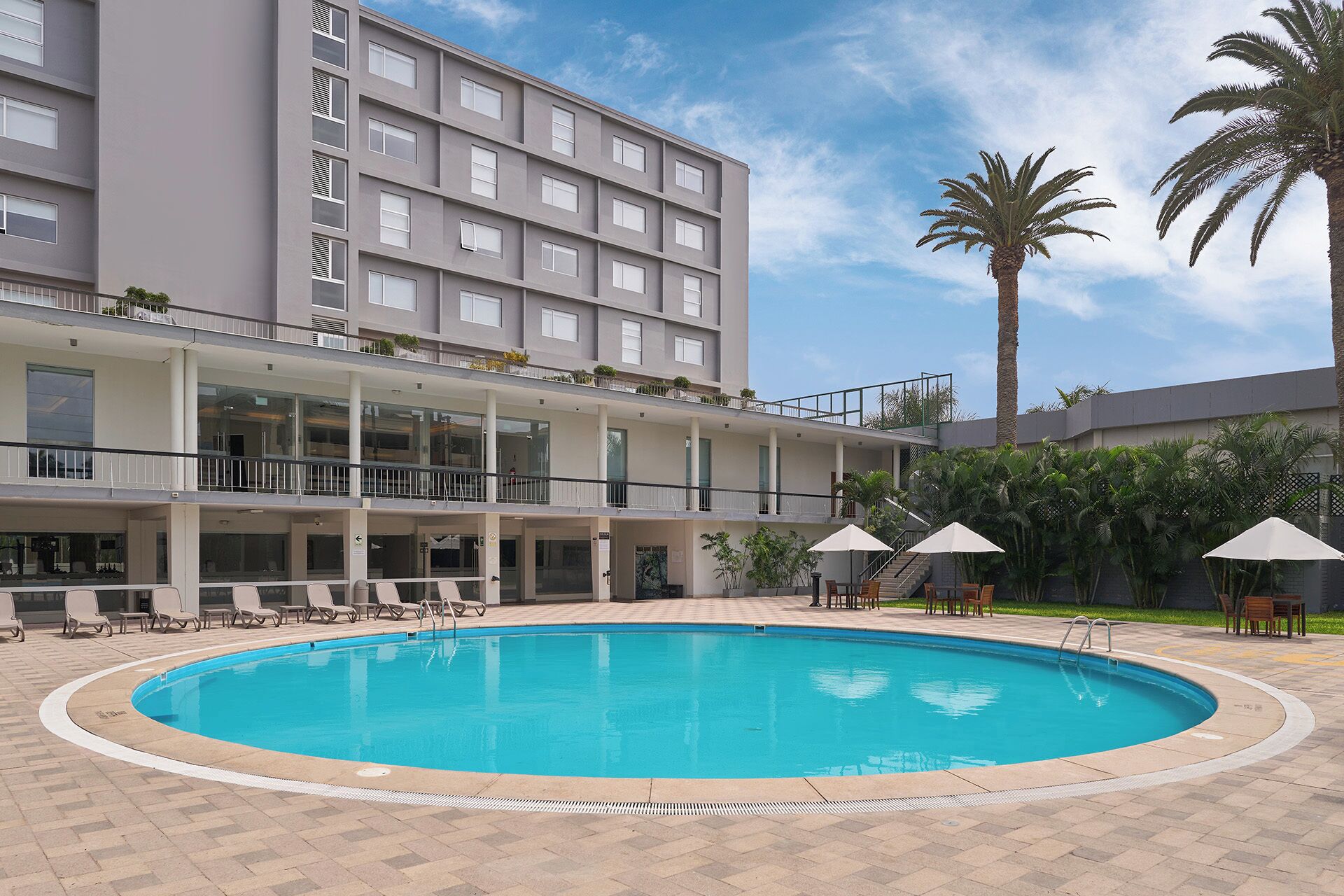 A swimming pool with lounge chairs and umbrellas, surrounded by a building with balconies and palm trees.