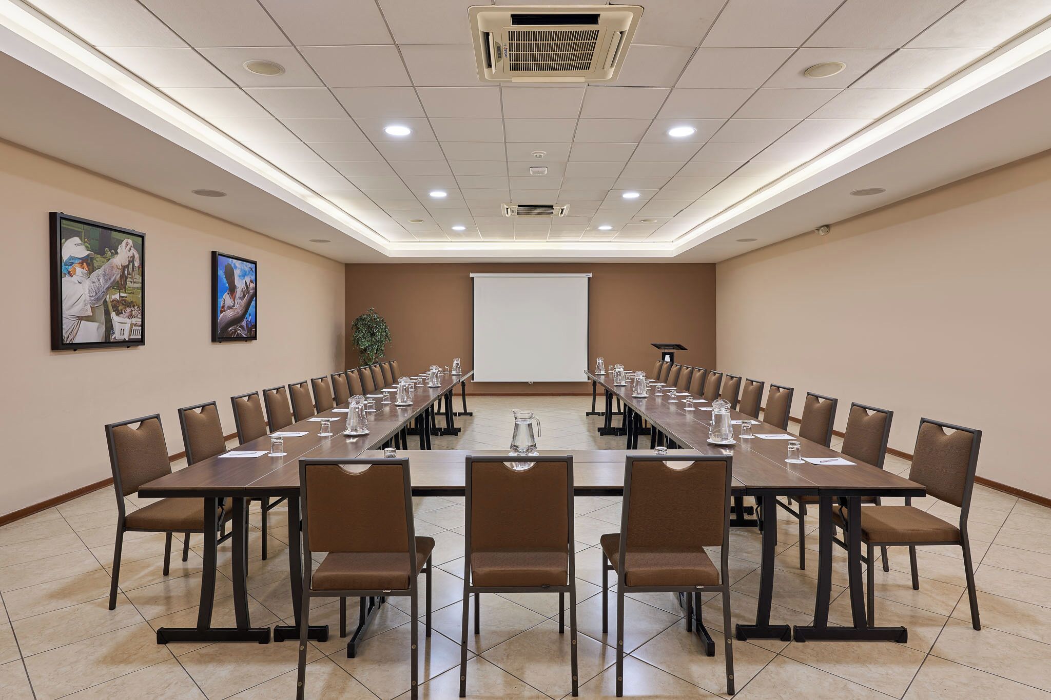 Un salón de conferencias con una larga mesa, sillas, una pantalla de proyector y una planta.