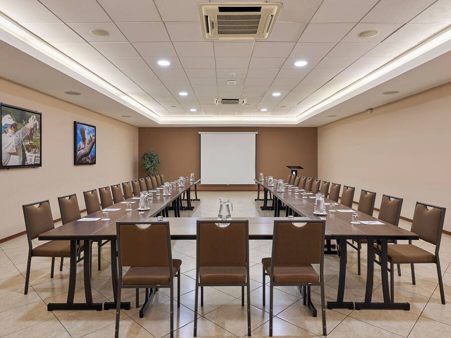 Un salón de conferencias con una larga mesa, sillas, una pantalla de proyector y una planta.