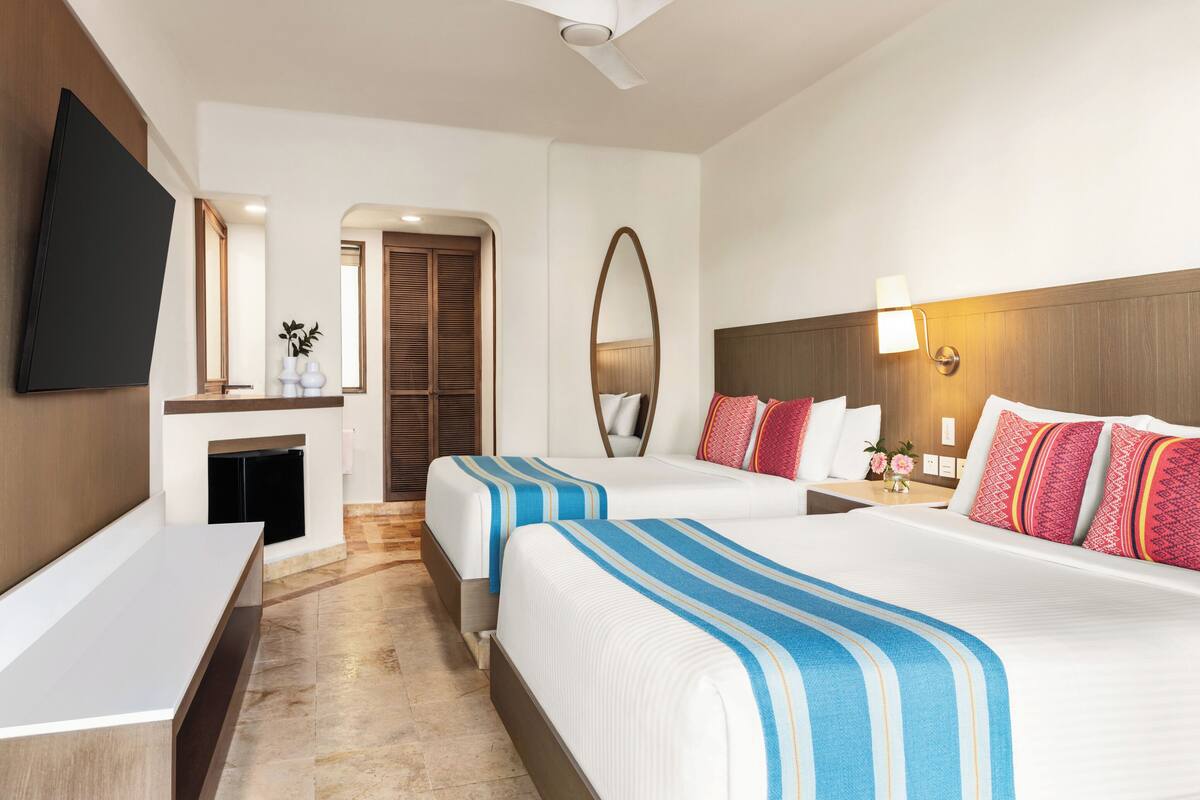 Habitación de hotel con dos camas, televisión de pantalla plana, escritorio, espejo y mesita de noche con lámpara.