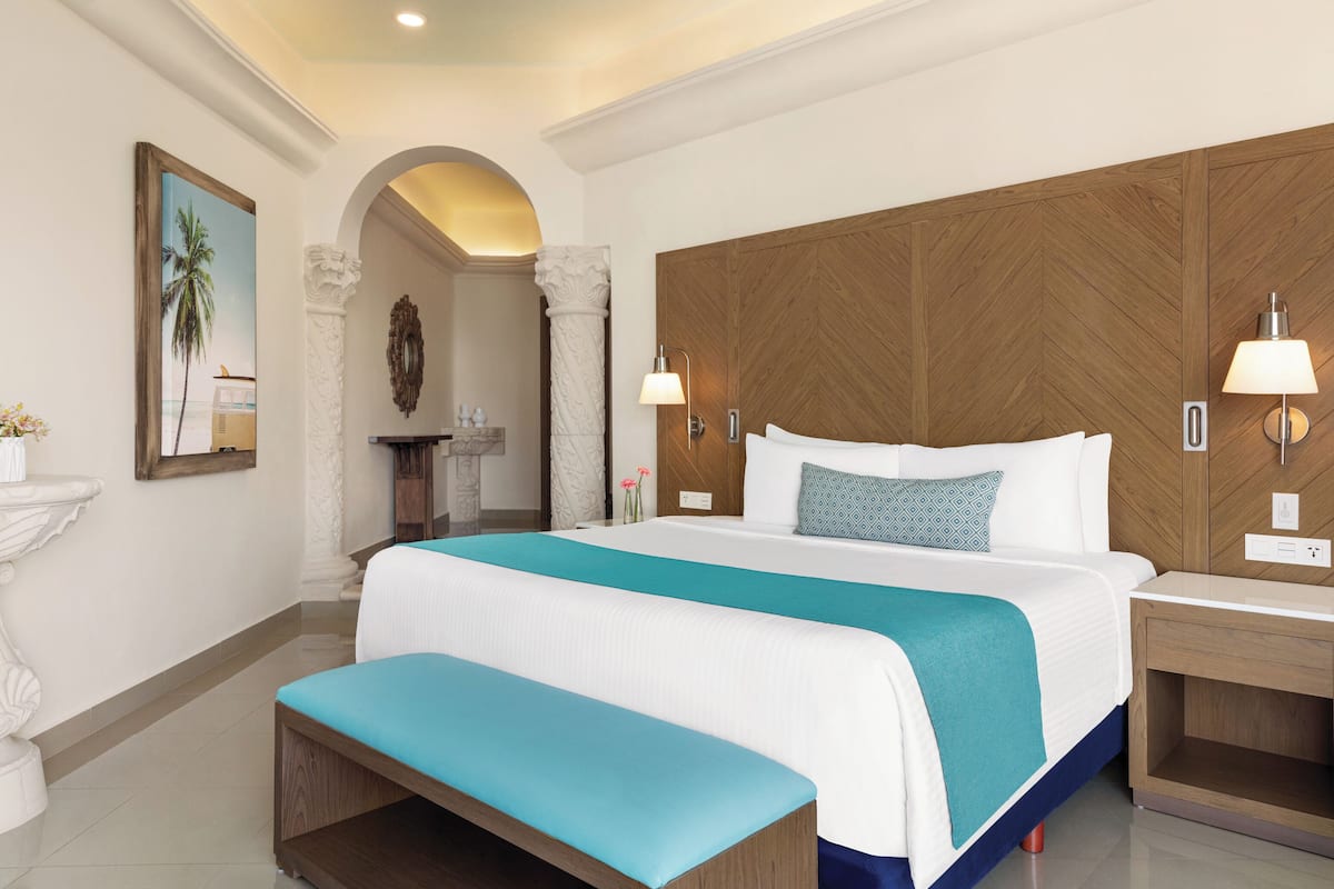 Una habitación de hotel con una cama grande, un banco, un cabecero de madera y una mesita de noche con una lámpara.