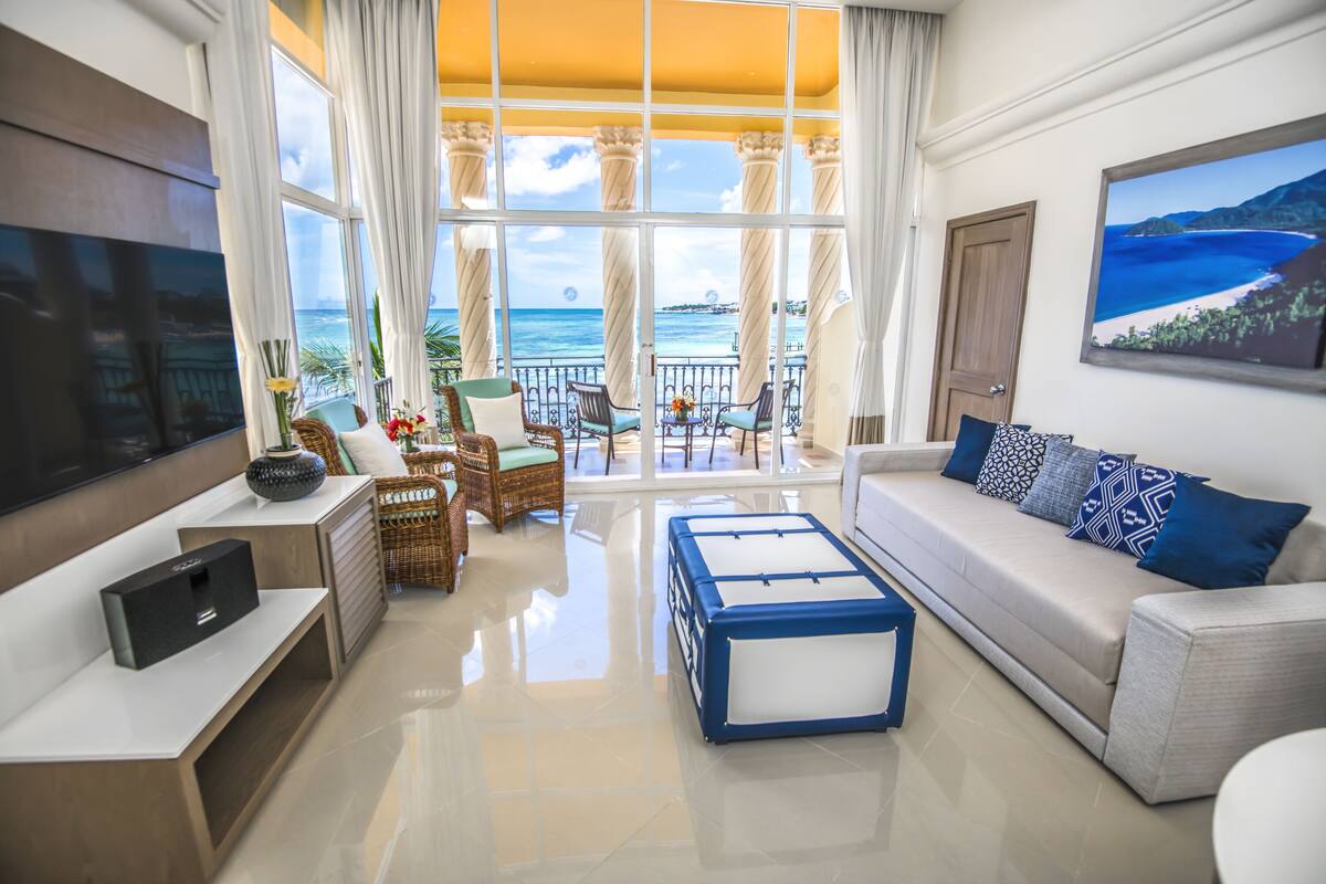 A modern living room with a large TV, comfortable seating, a balcony with ocean views, and a painting on the wall.