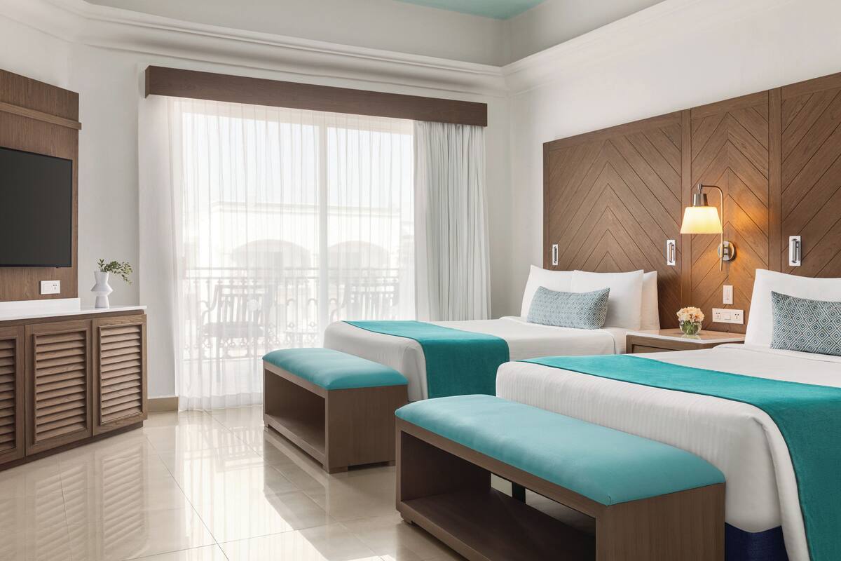 Habitación de hotel con dos camas, televisión, cabecero de madera, ventana con cortinas translúcidas y una mesita con un florero.
