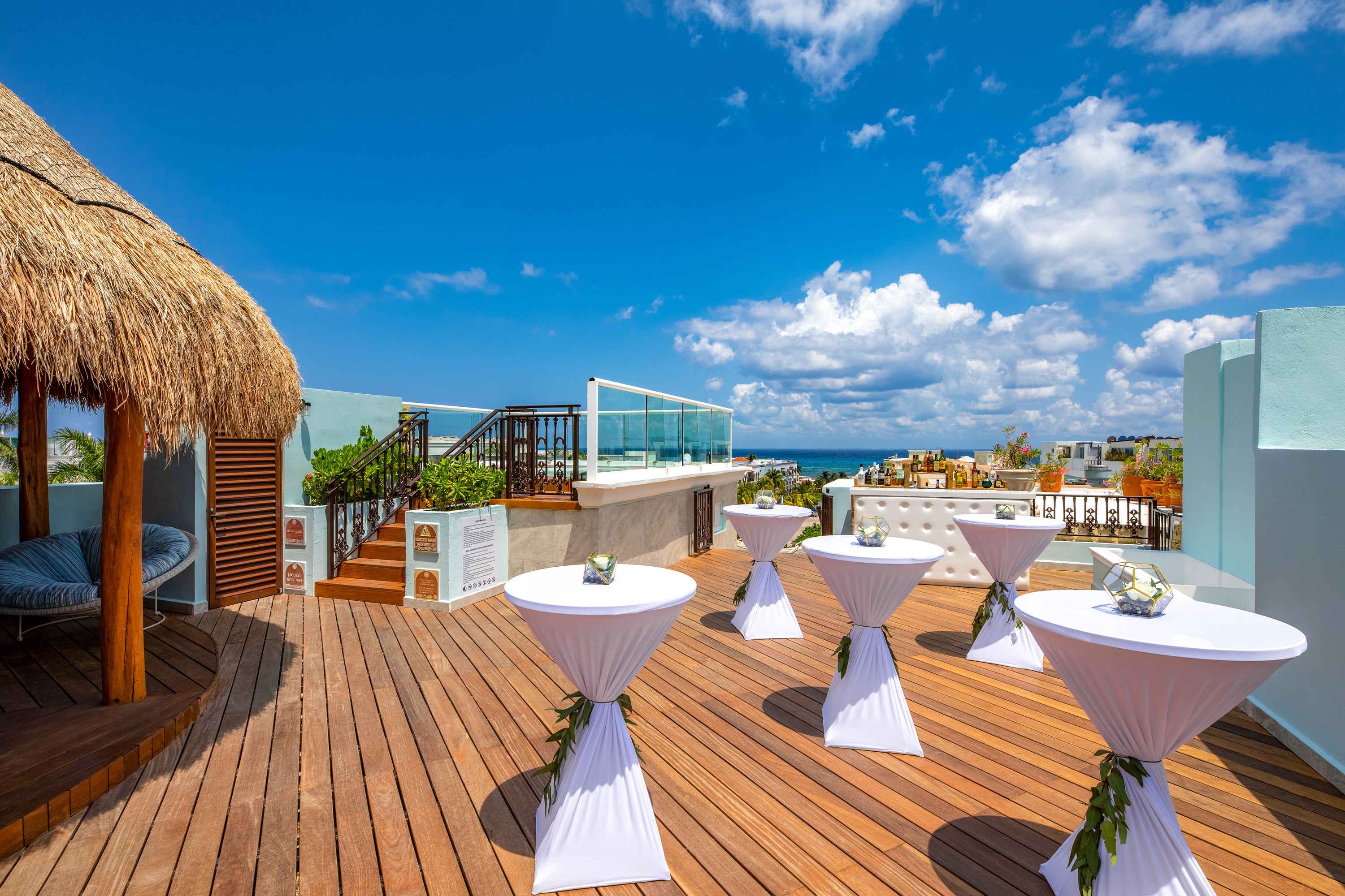 A wooden deck with white tables and chairs, a thatched roof structure, and a glass balustrade overlooking the ocean.