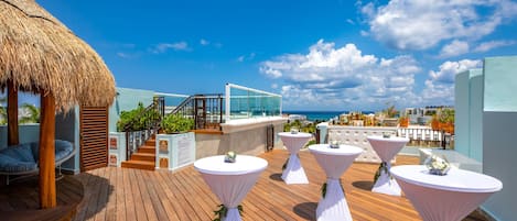 A wooden deck with white tables and chairs, a thatched roof structure, and a glass balustrade overlooking the ocean.