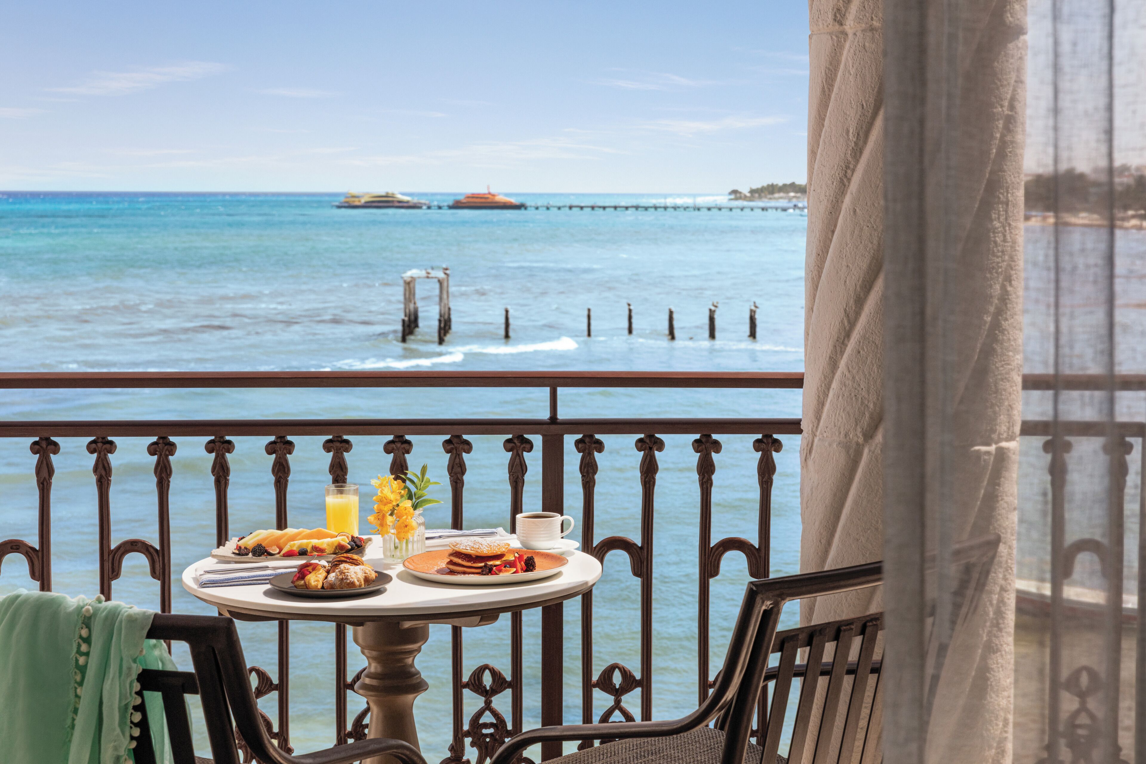 A table with breakfast items and a glass of orange juice, overlooking a calm sea with a pier in the distance.