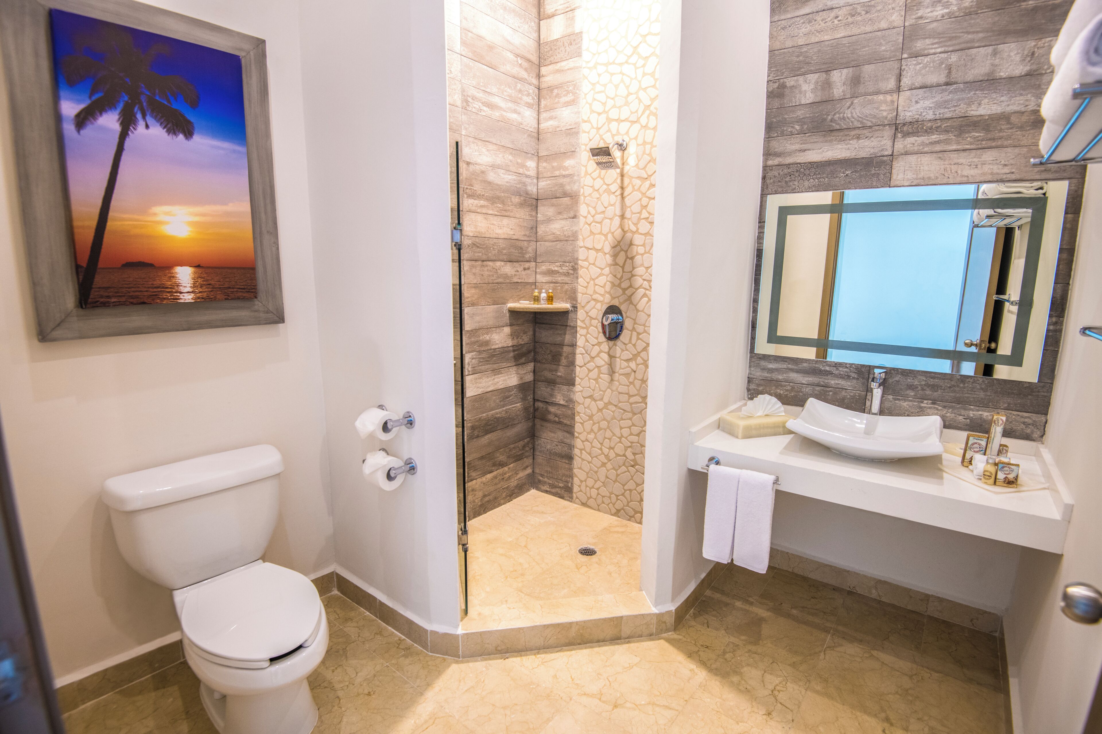 Une salle de bain comprenant des toilettes, une douche avec une porte en verre, un lavabo avec un miroir et un tableau représentant un coucher de soleil.
