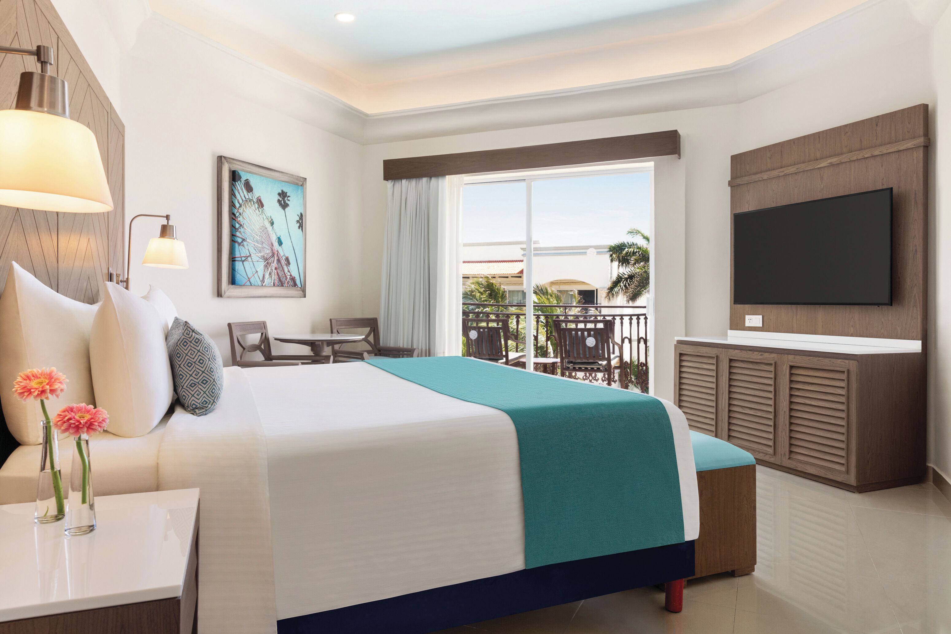 Ein Hotelzimmer mit einem großen Bett, einem Schreibtisch mit Lampe, einem an der Wand montierten Fernseher und einem Balkon mit Sitzmöbeln.