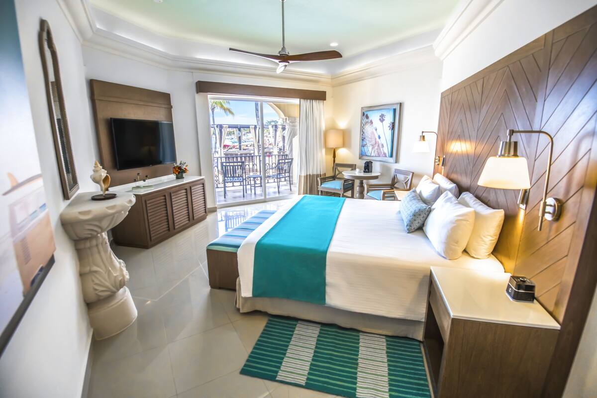 Una habitación de hotel moderna con una cama grande, televisión, balcón con vistas y escritorio con lámpara.