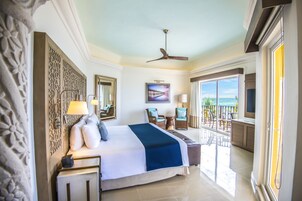 A hotel room with a large bed, a balcony with a view of the ocean, a ceiling fan, and a painting on the wall.