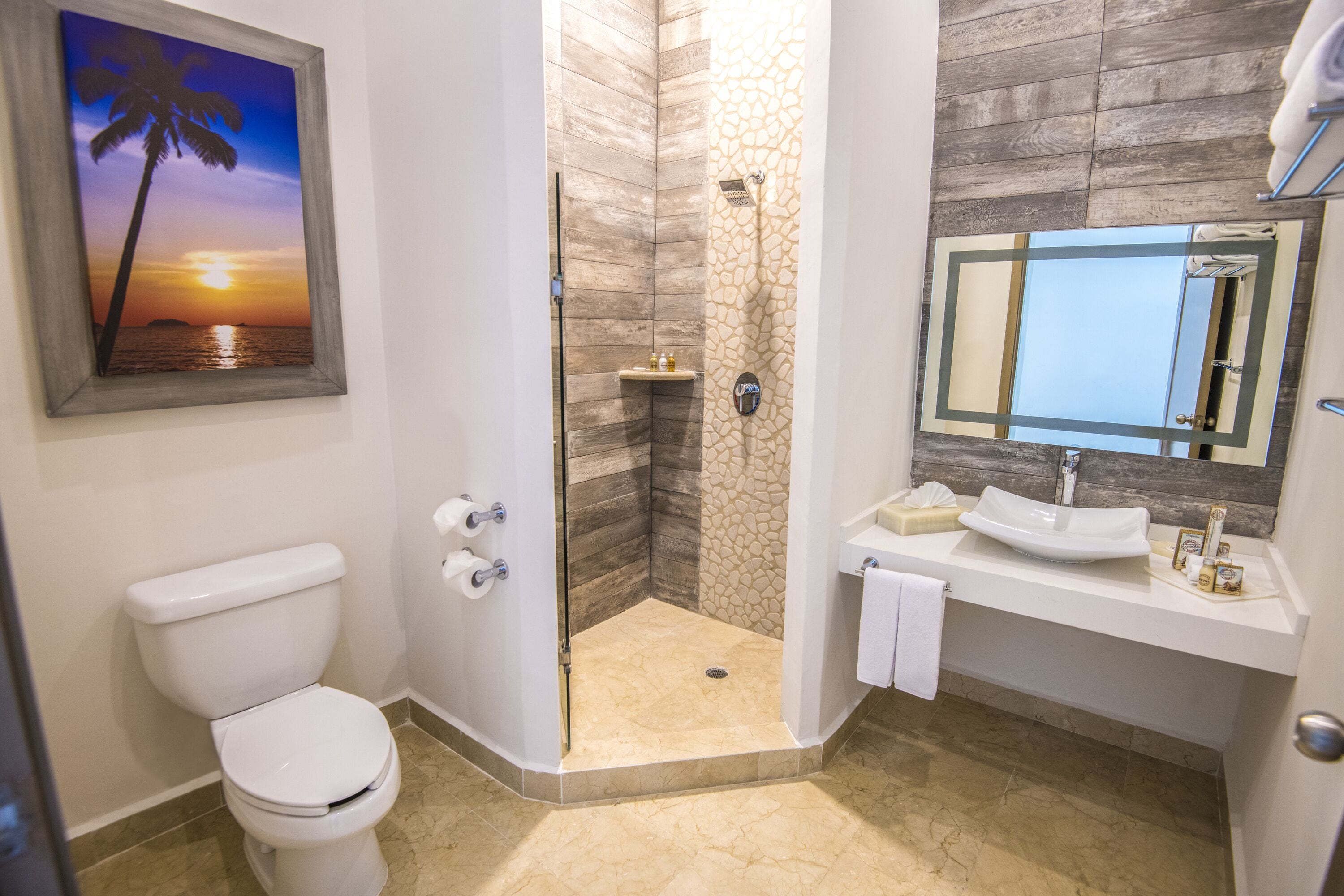 A bathroom with a toilet, a shower with a stone wall, a sink with a mirror, and a sunset painting on the wall.