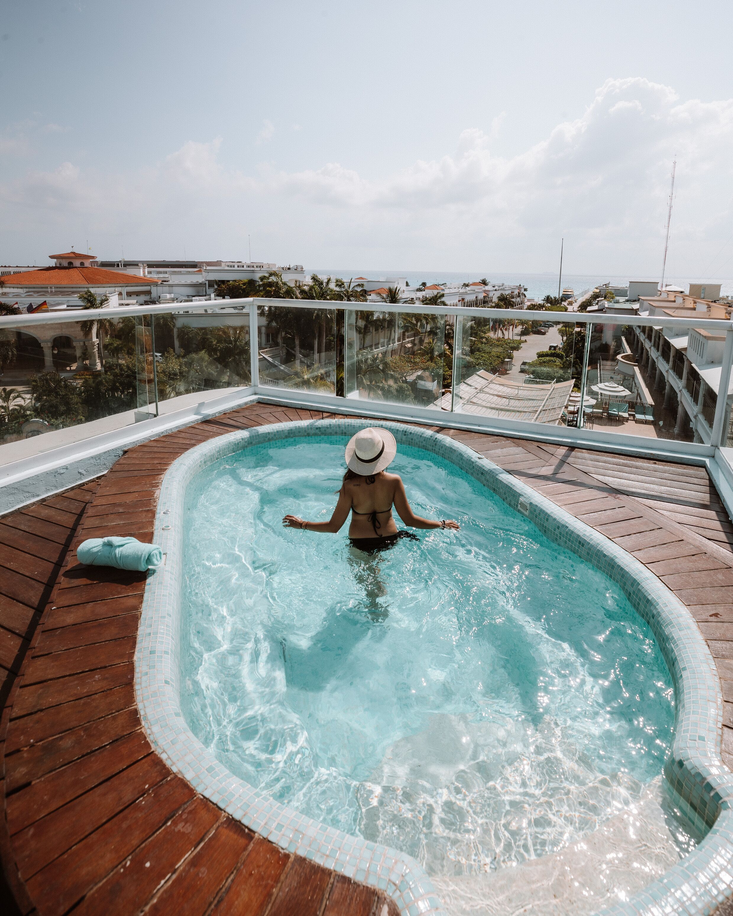 Eine Person in einem Schwimmbecken auf einer Holzterrasse mit Blick auf das Meer und Gebäude im Hintergrund.