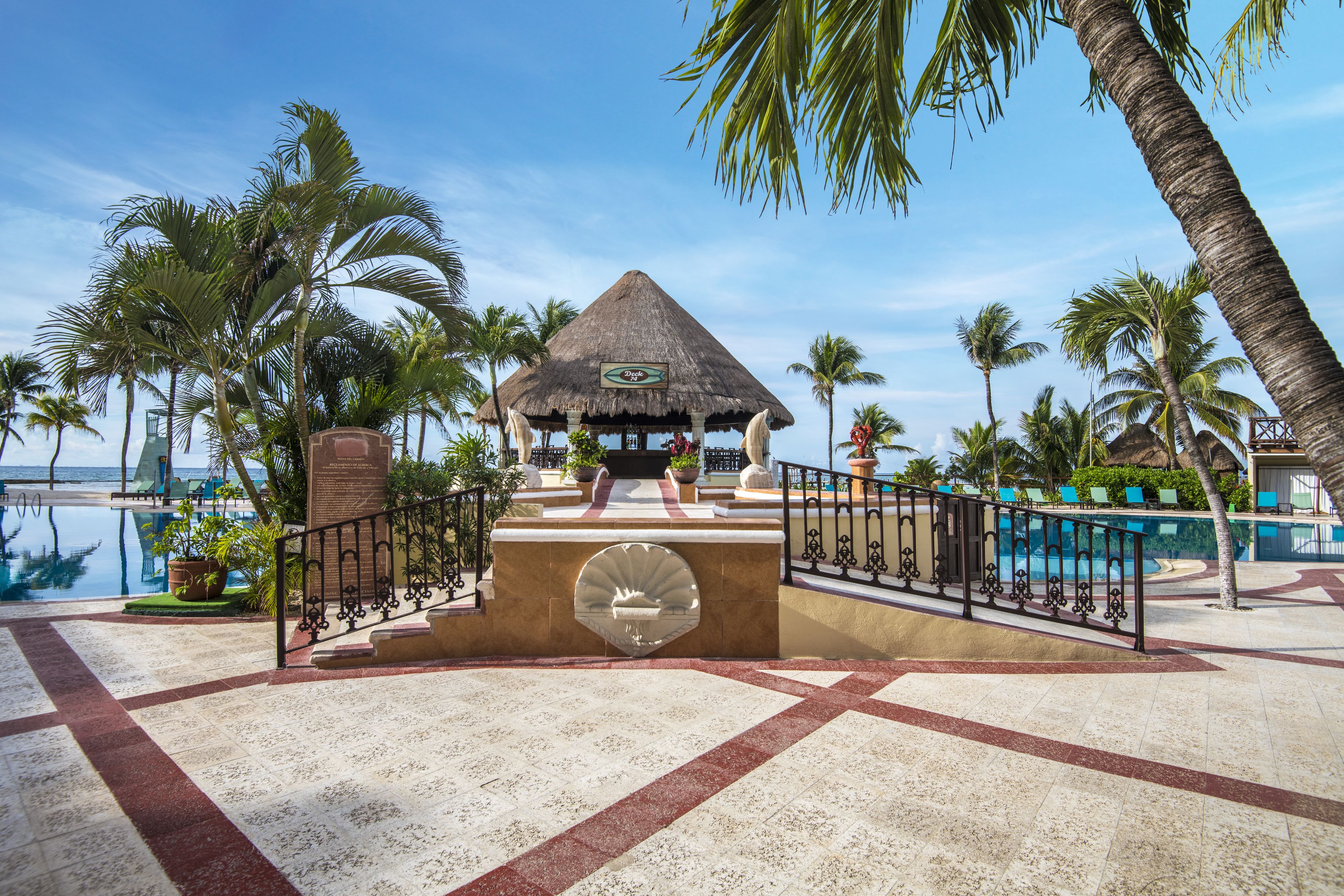 A resort pool area with a thatched-roof gazebo, palm trees, and a clear blue sky.