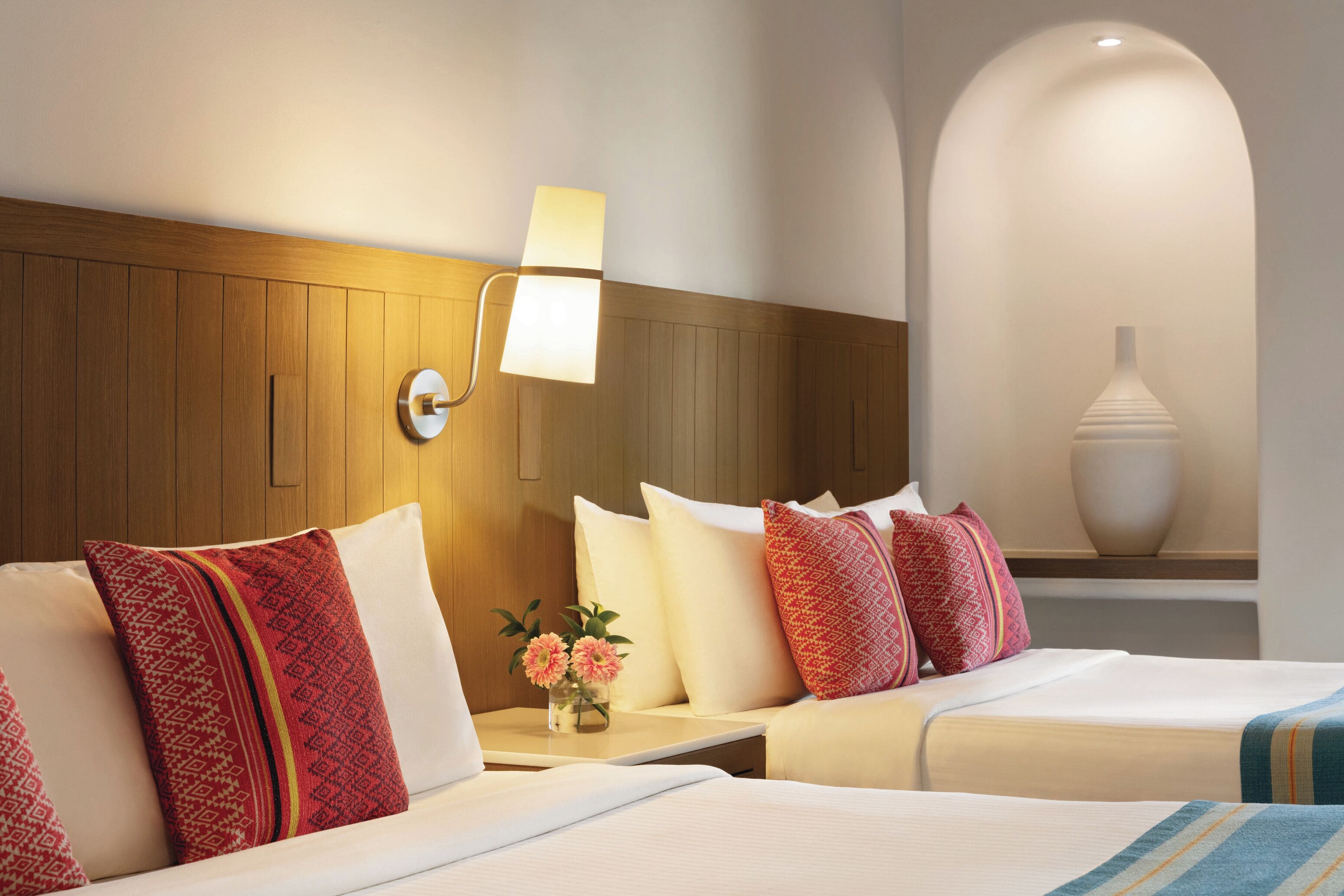 A hotel room with two beds, a wooden headboard, a nightstand with a lamp, and a vase on a shelf.