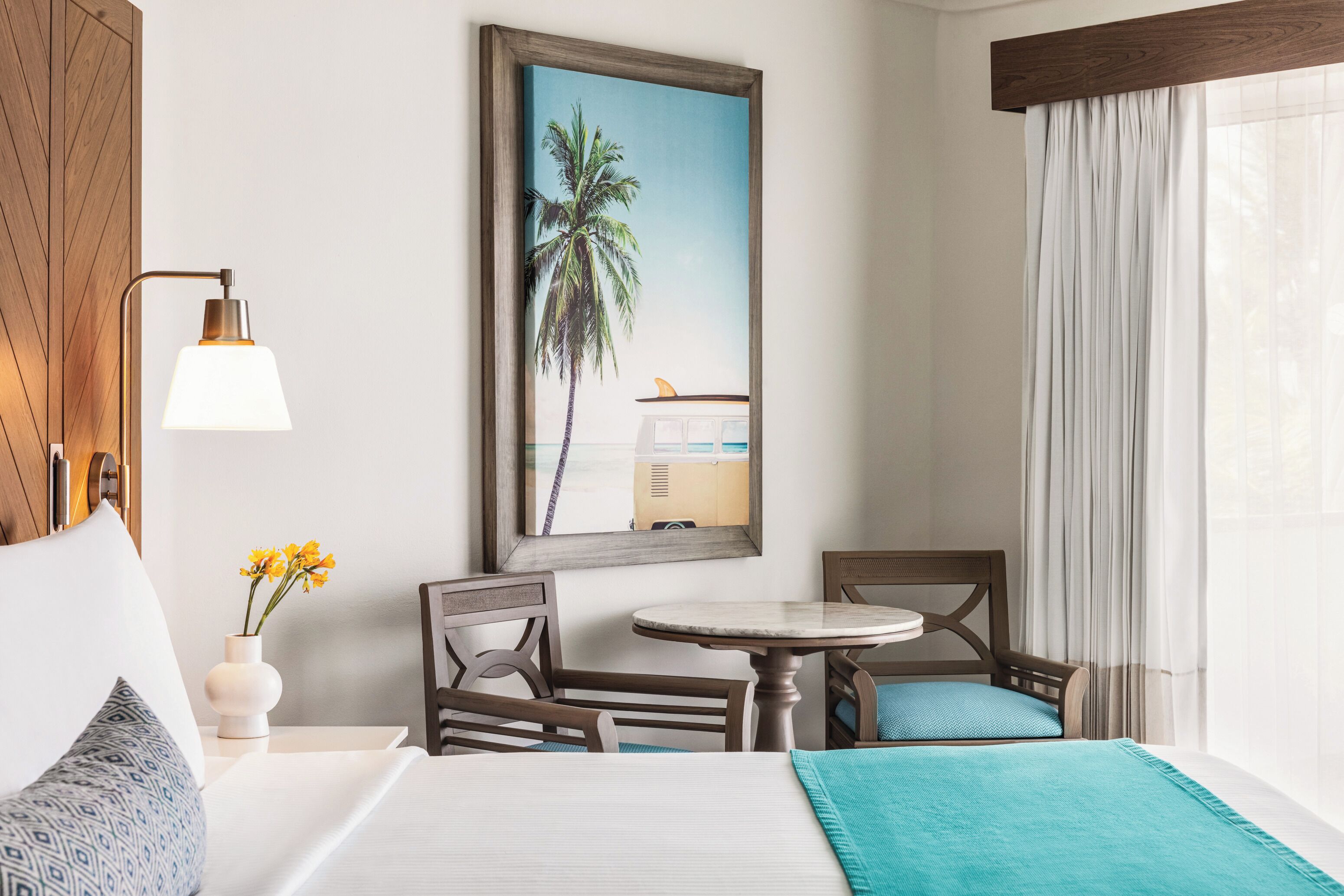 A hotel room with a bed, a small table, two chairs, a nightstand with a lamp, a painting of a palm tree, and a window with curtains.