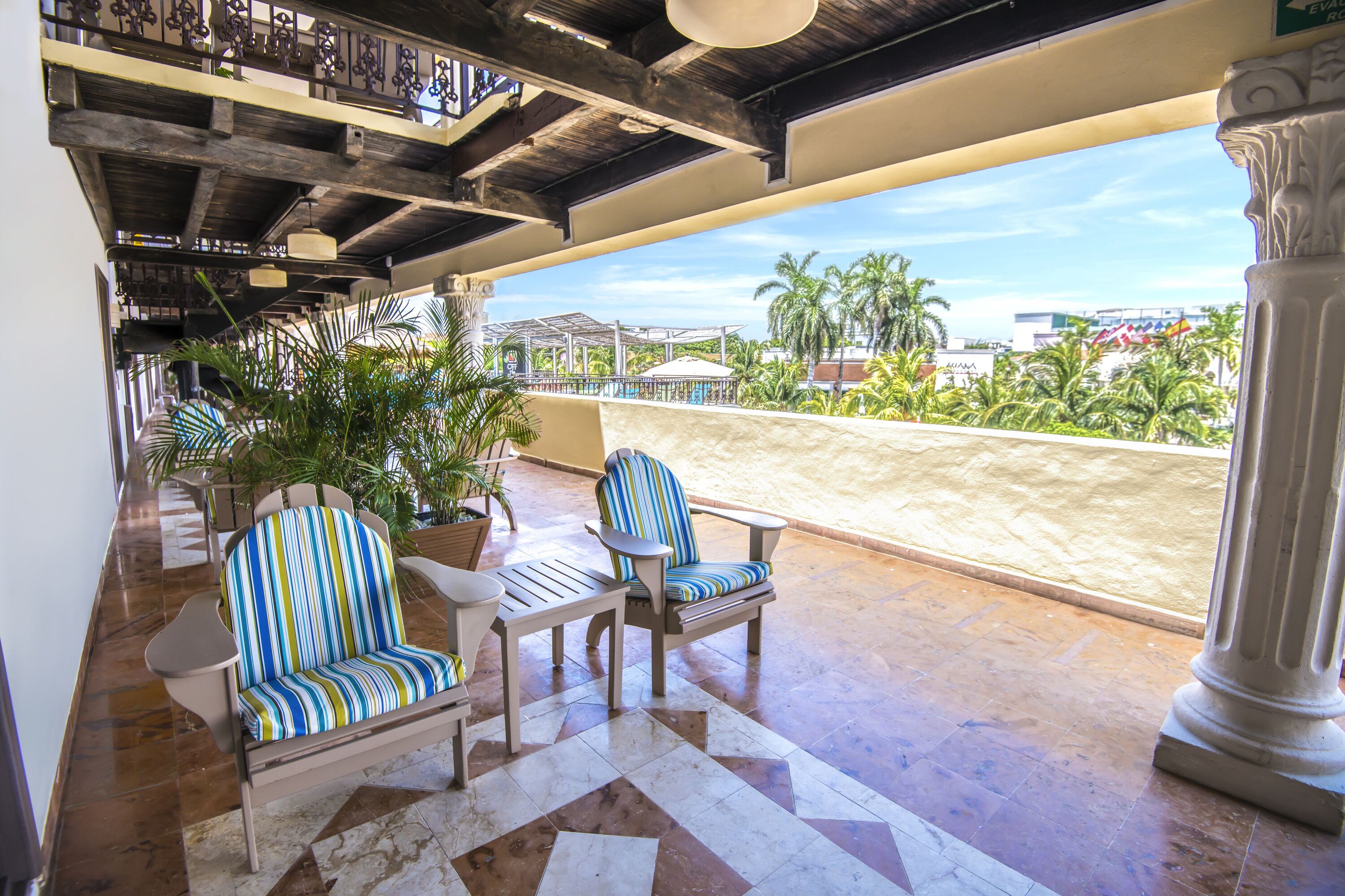 A covered outdoor seating area with two striped chairs and a table, a potted plant, and a view of palm trees and buildings in the background.