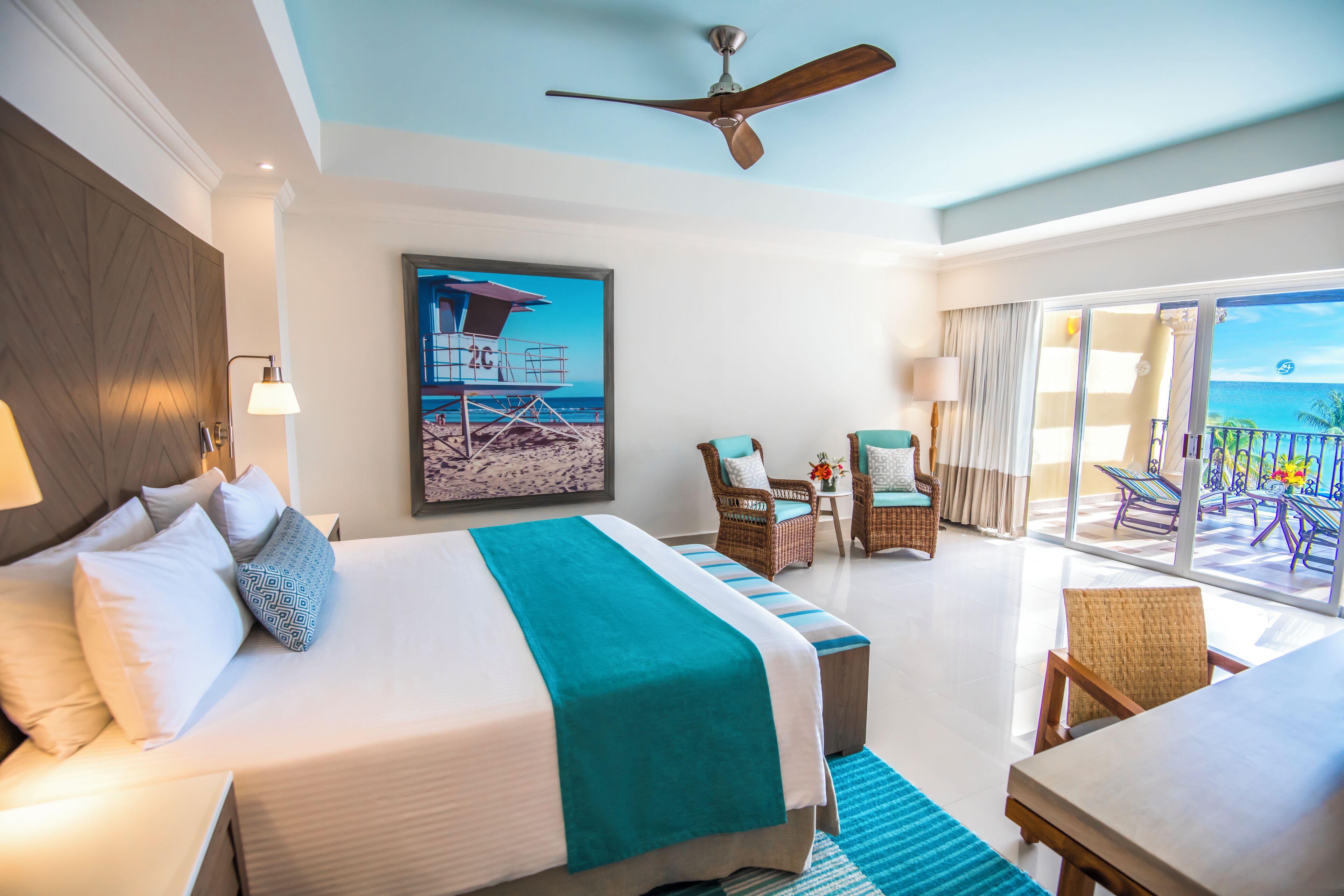 A hotel room with a bed, a ceiling fan, a balcony with chairs, and a view of the ocean.