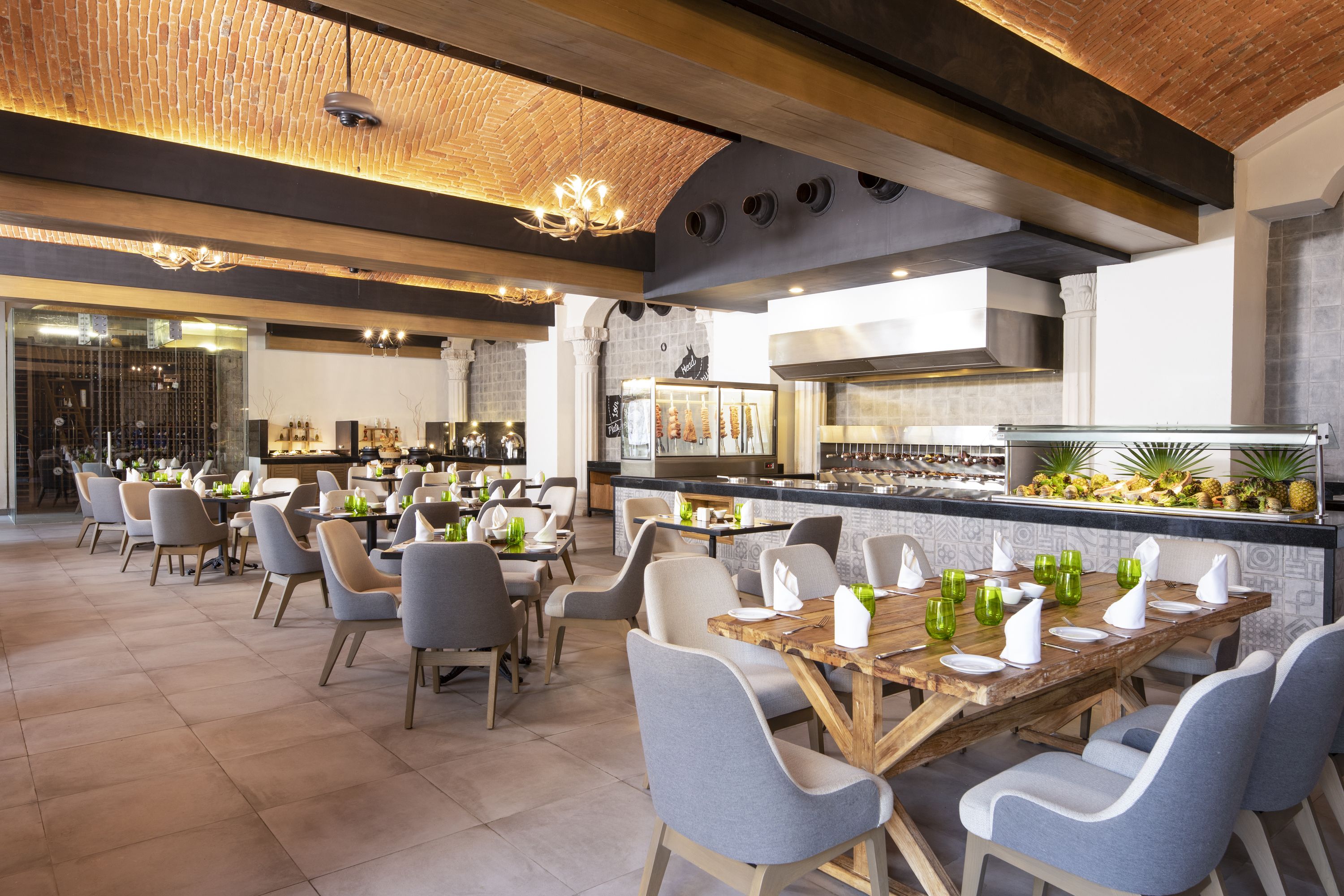 A spacious restaurant with high ceilings, exposed brick, and modern lighting. Tables are set with chairs and green glassware.