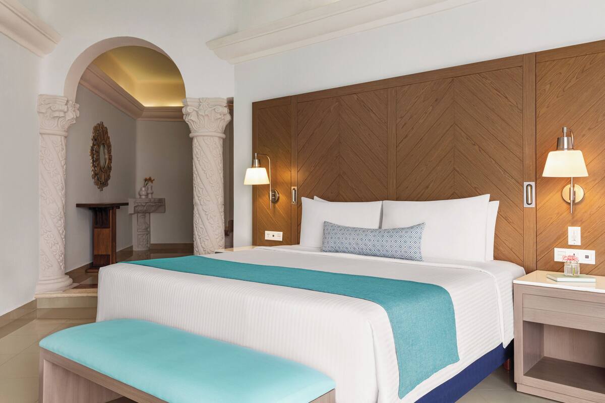A hotel room with a large bed, a bench, a wooden headboard, two bedside lamps, and a small table with a lamp.