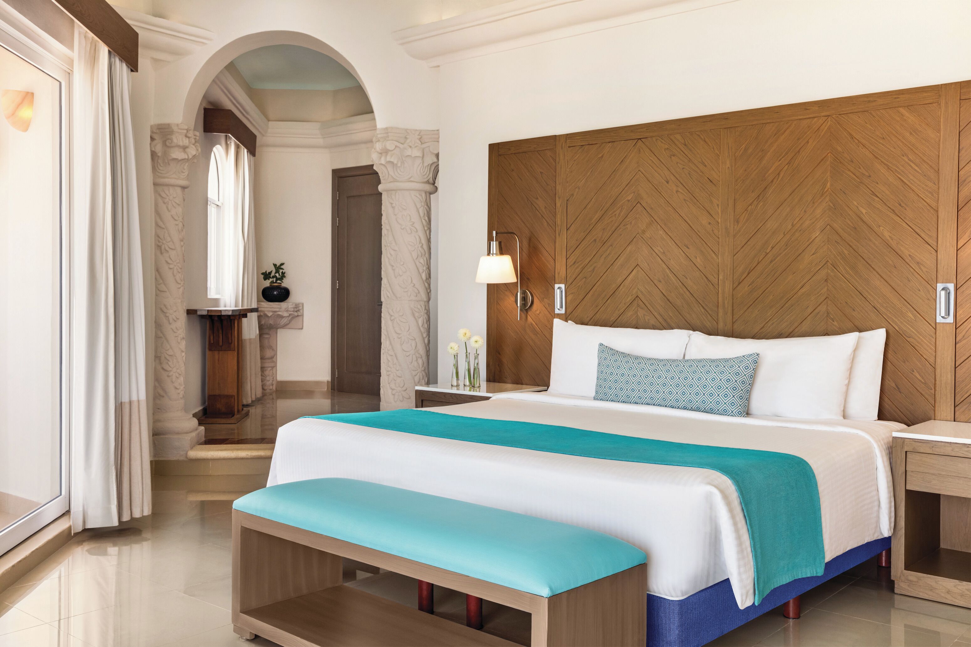 Habitación de hotel con una cama grande, cabecero de madera, un banco y una mesita de noche con lámpara.