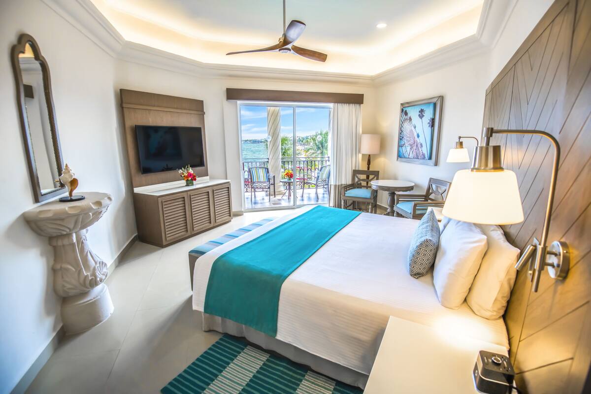 Habitación de hotel con cama, televisor, ventilador de techo, balcón con vistas y un cuadro en la pared.