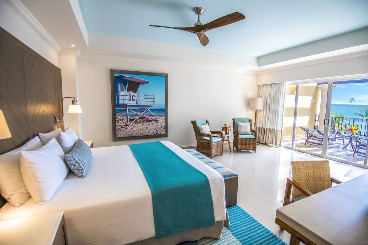 Habitación de hotel con cama, ventilador de techo, balcón con sillas y vista a la playa.