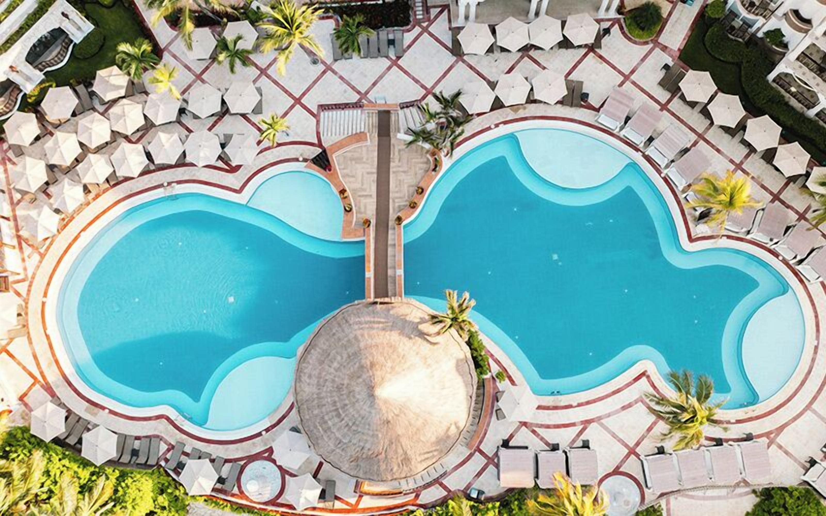 Vue aérienne d’une piscine divisée en deux zones distinctes, entourée de chaises longues et de palmiers.