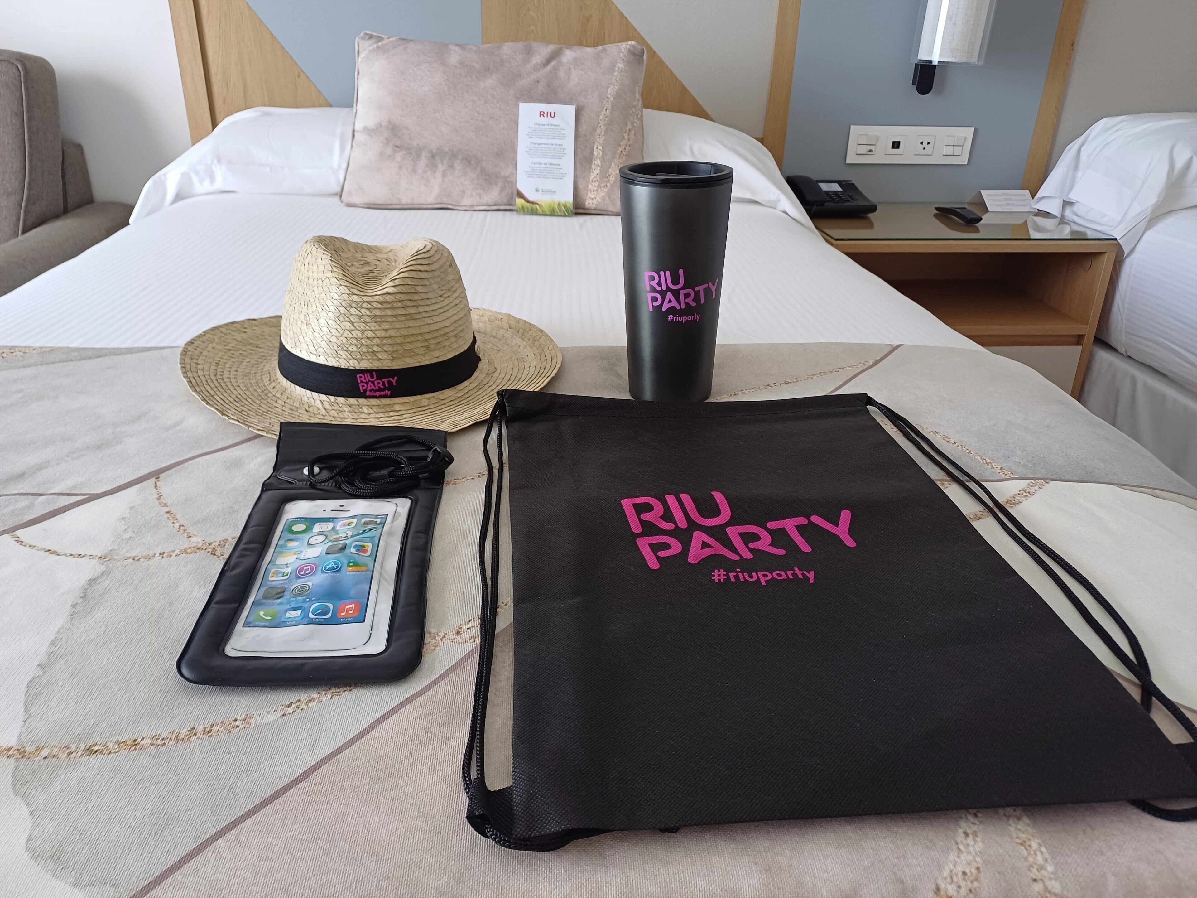Una cama bien hecha con un almohadón, un sombrero de paja, un vaso negro, una bolsa de tela negra con cordón y un teléfono inteligente sobre la colcha.