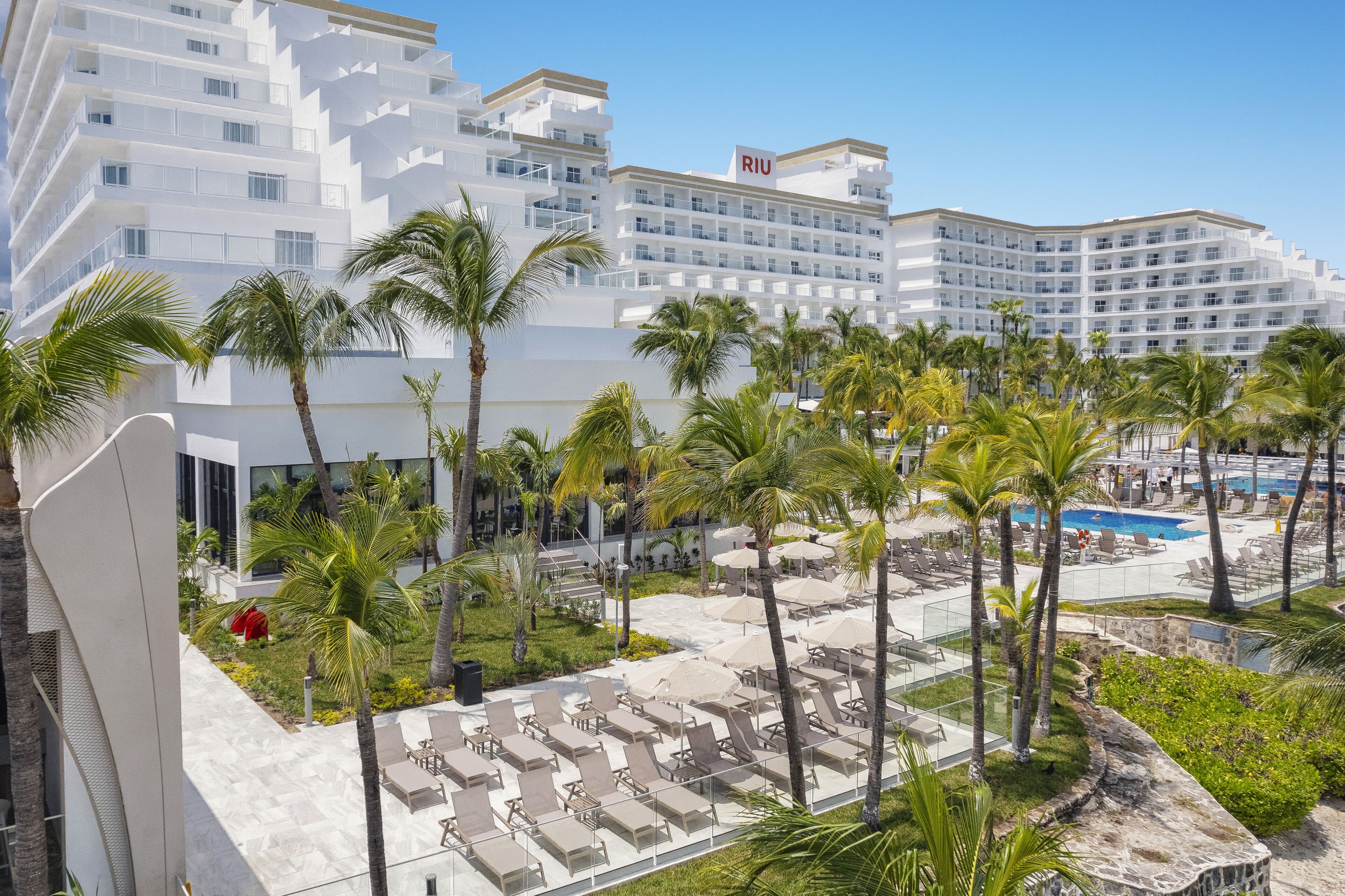 A resort with multiple white buildings, a pool area with sun loungers, and palm trees.