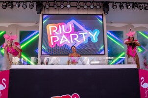 Un escenario con una pantalla grande que muestra 'RIU PARTY' y artistas con trajes rosas.
