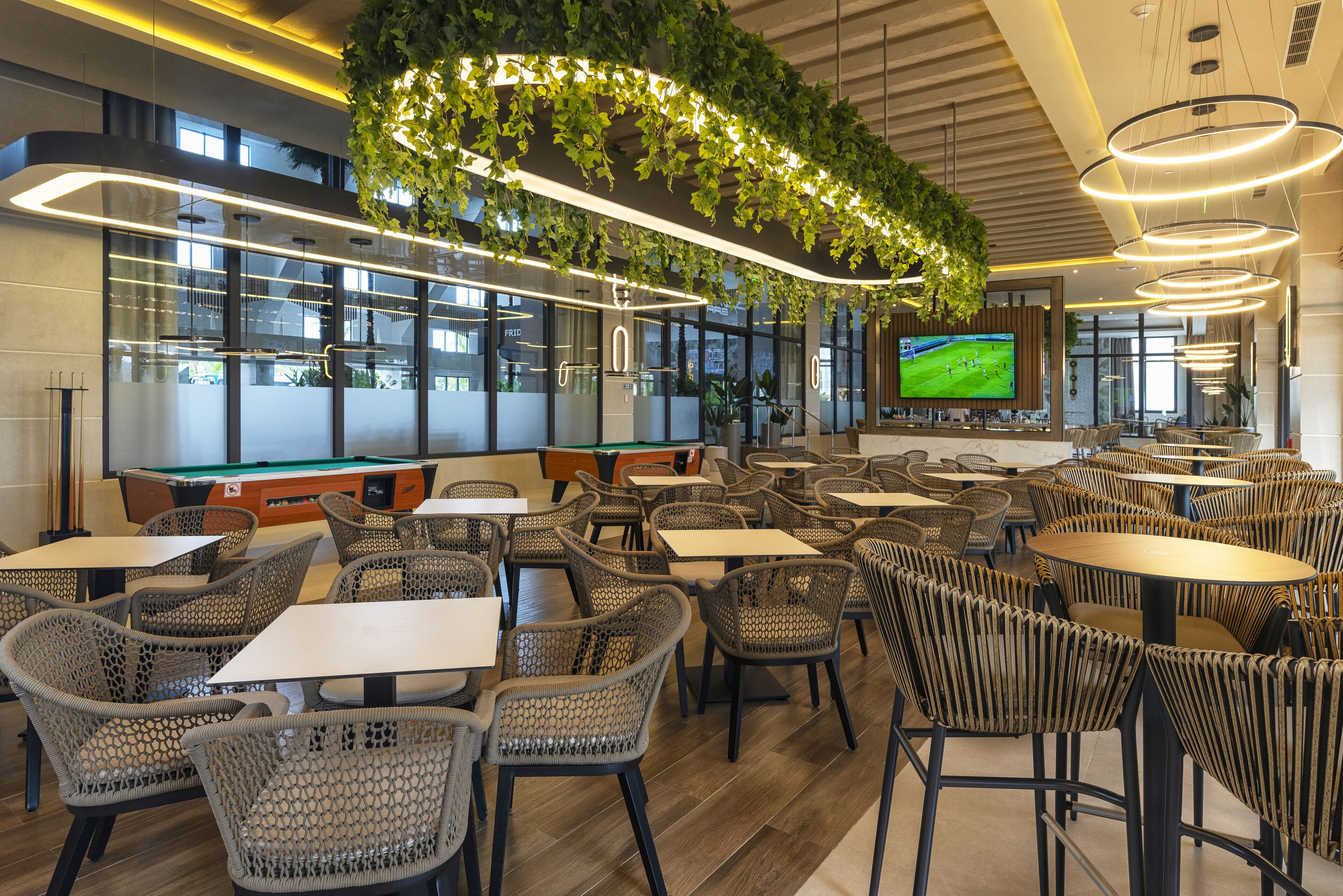 A modern indoor dining area with wicker chairs, rectangular tables, and a large TV screen displaying a sports game.