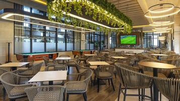 A modern indoor dining area with wicker chairs, rectangular tables, and a large TV screen displaying a sports game.