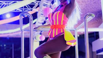 Una persona vestida con un atuendo de colores brillantes, de pie en una plataforma con barandas metálicas, rodeada de gente.