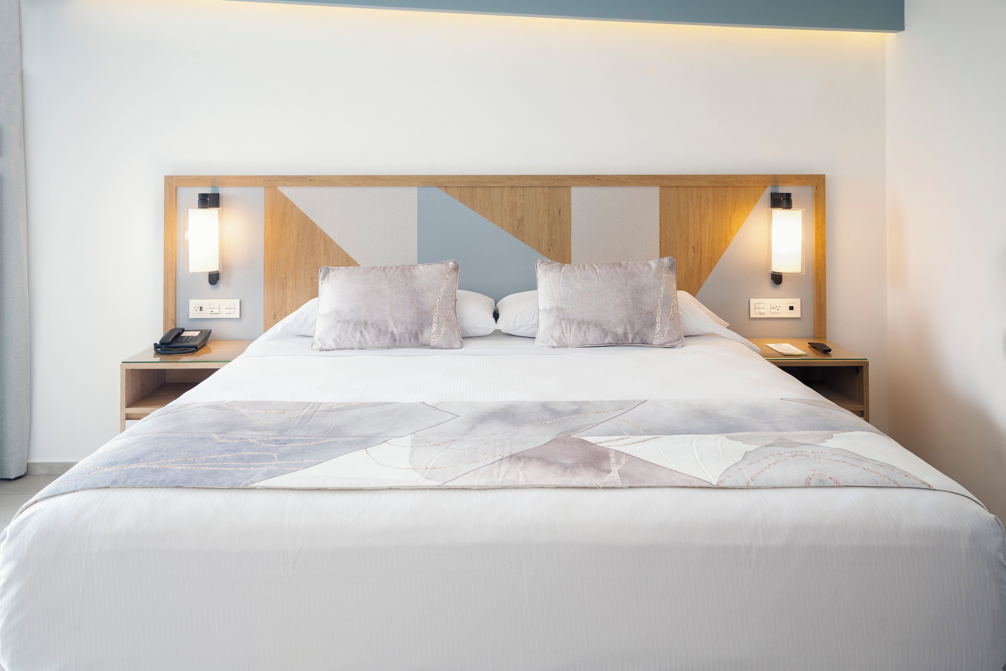 Un lit bien fait, avec une couvre-lit à motifs géométriques, deux oreillers blancs et une tête de lit en bois agrémentée de lampes fixées au mur.