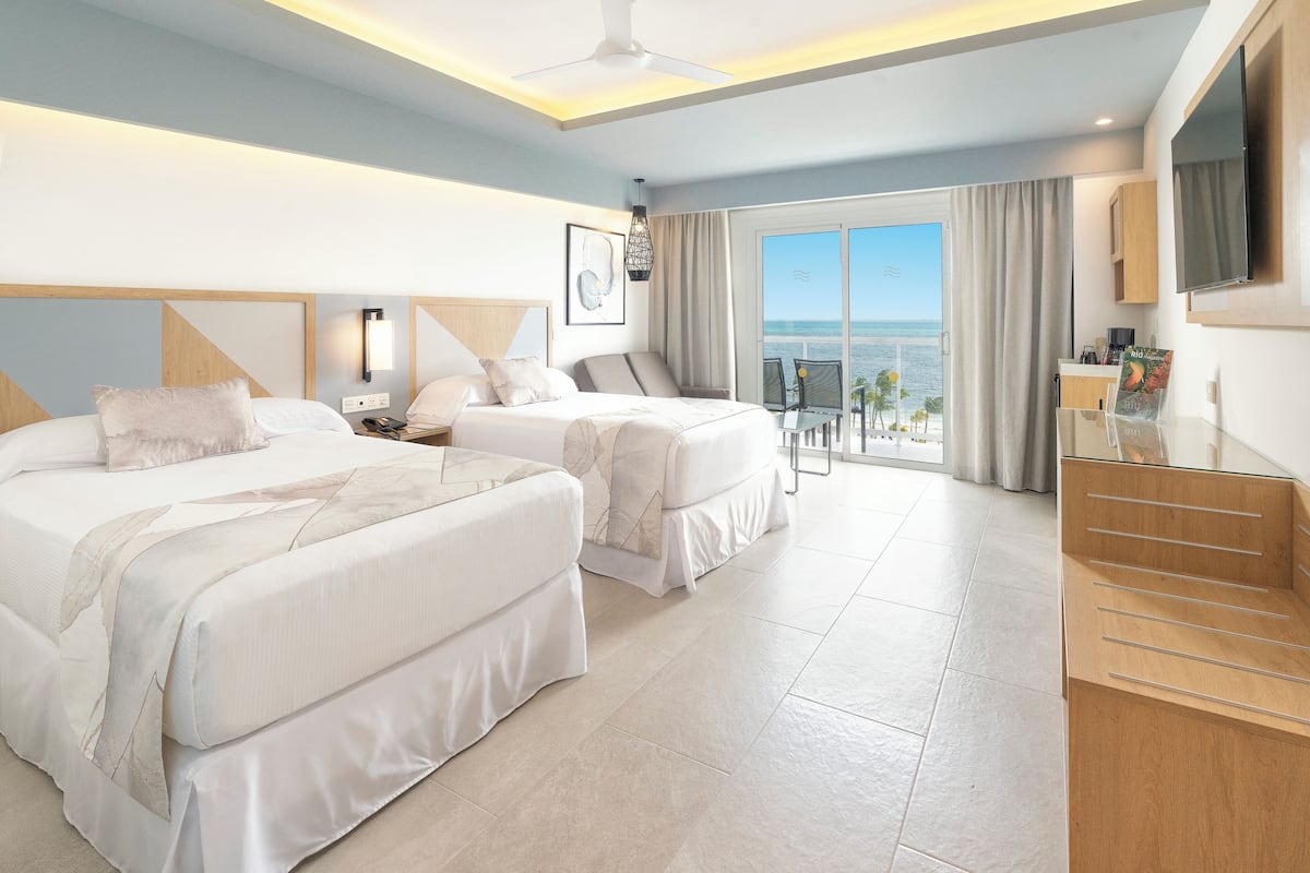 Habitación de hotel con dos camas, un balcón con vista al mar, un televisor de pantalla plana y un escritorio de madera.
