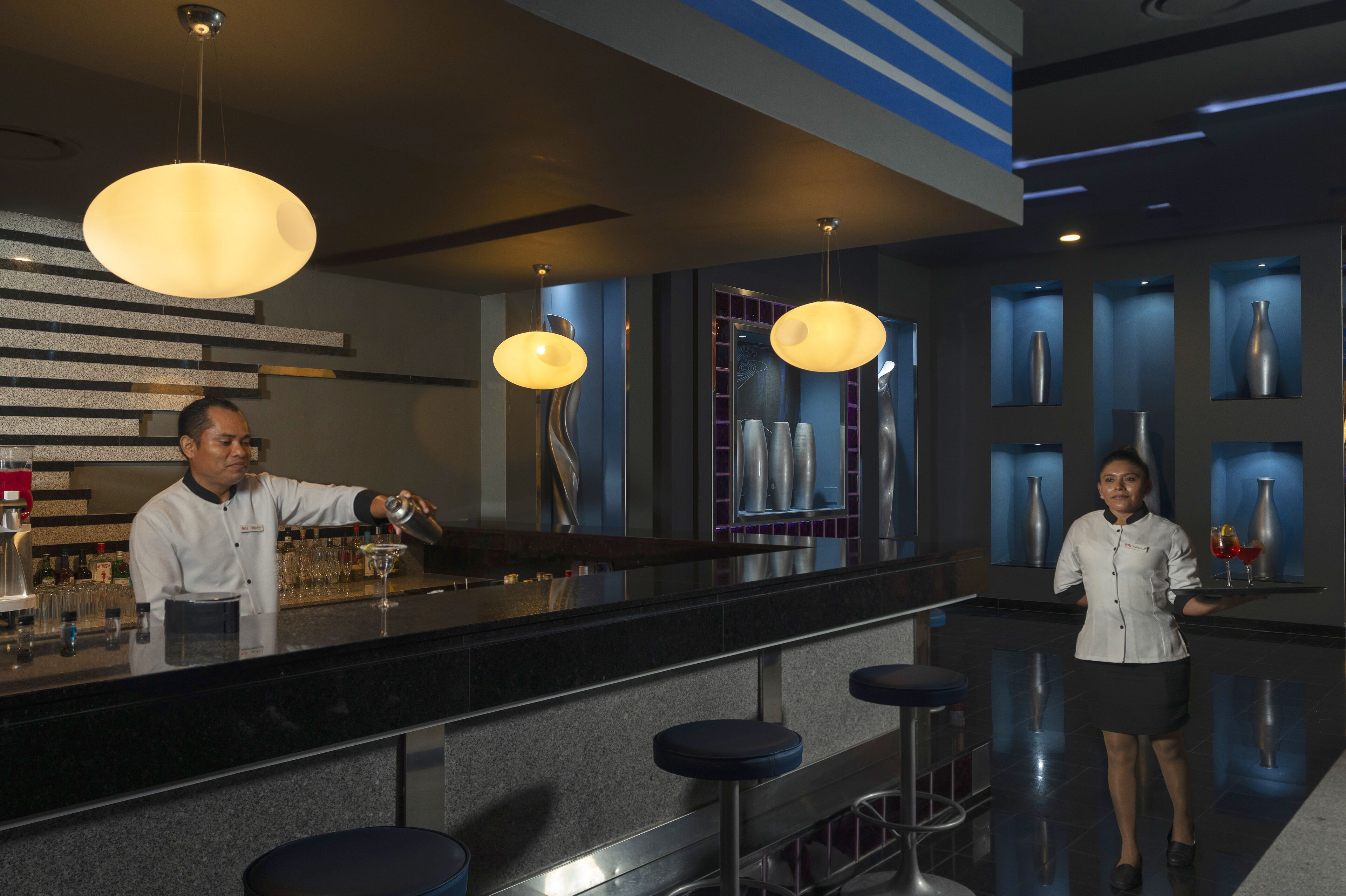 Zona bar moderna con un barista che prepara un cocktail, una donna che serve e un design interno elegante e contemporaneo.