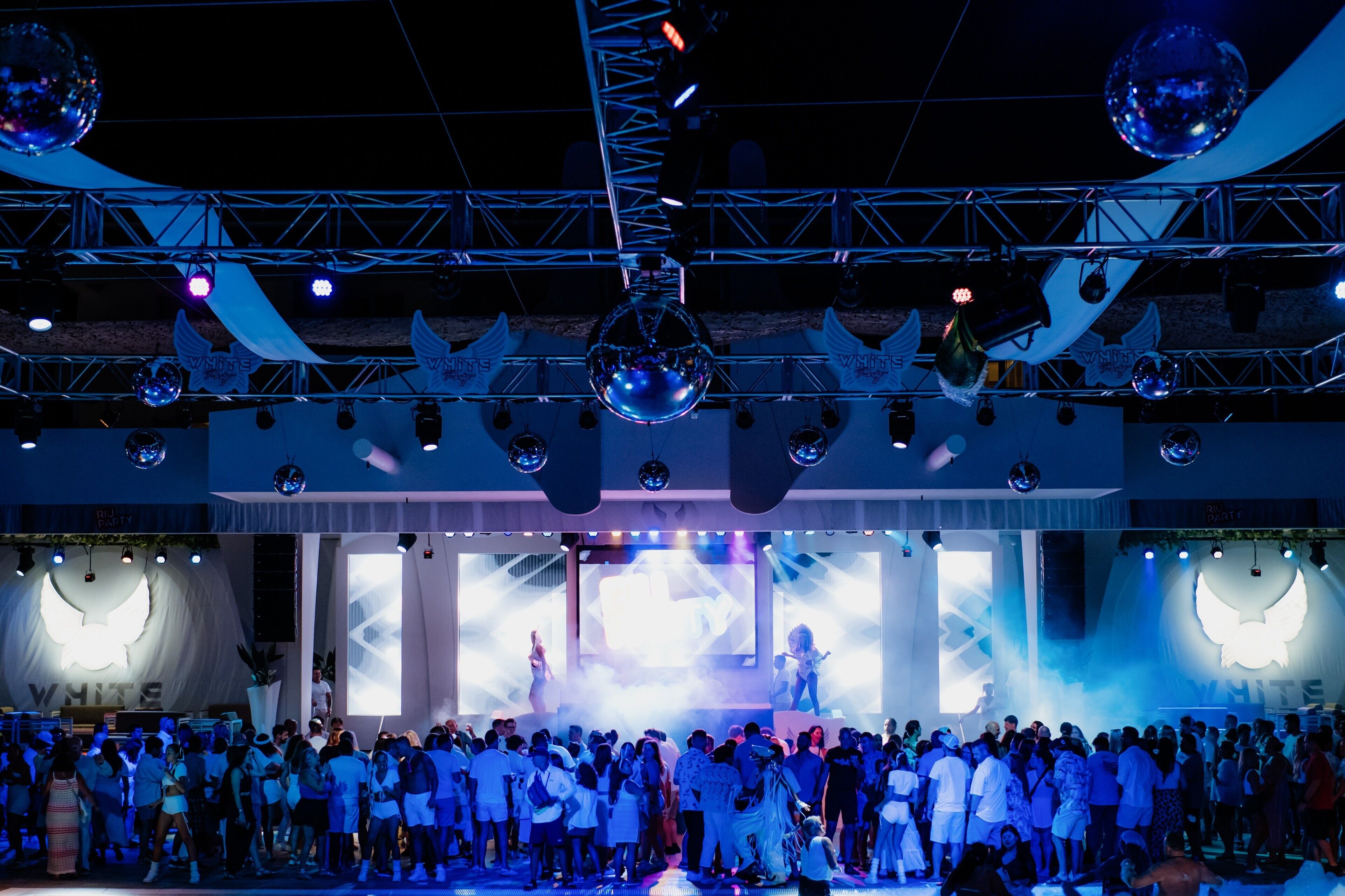 Un evento multitudinario en interior con un escenario, bolas de discoteca y una pantalla grande.