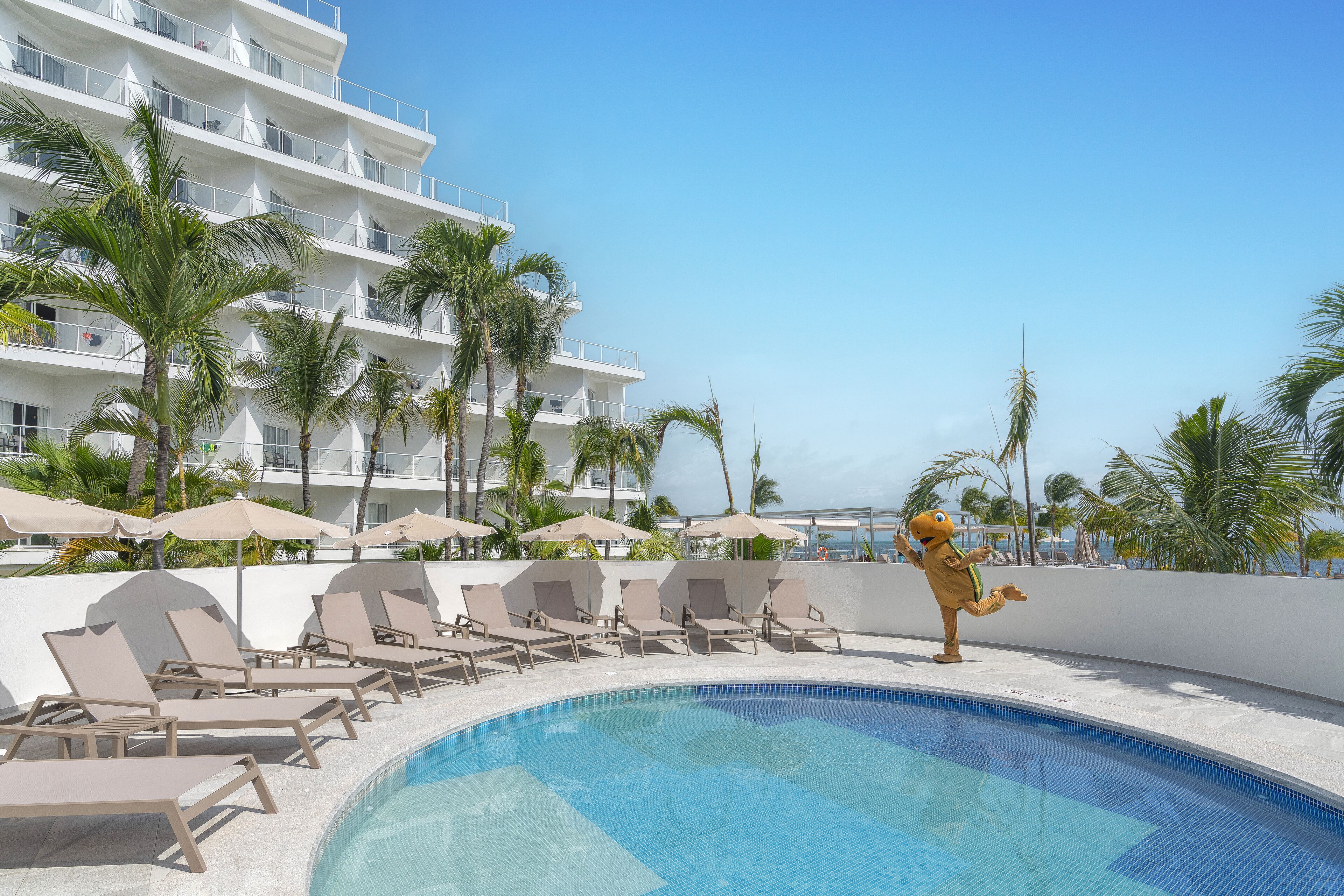 A poolside area with lounge chairs and umbrellas, a building with balconies, and palm trees.