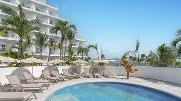 A poolside area with lounge chairs and umbrellas, a building with balconies, and palm trees.