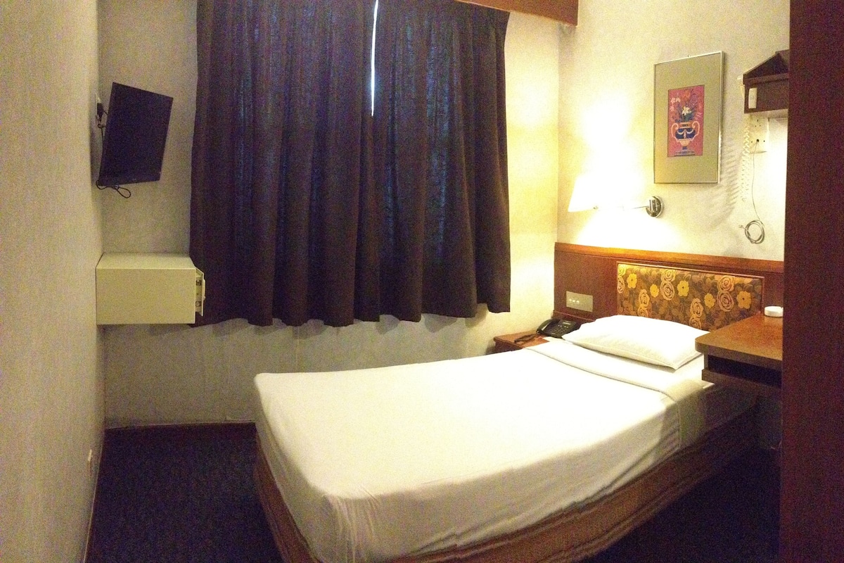 A hotel room with a single bed, a bedside table, a nightstand, a wall-mounted TV, and a curtain.