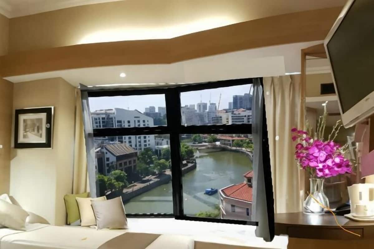 A hotel room with a large window, a bed, a desk with a computer, and a vase of flowers.