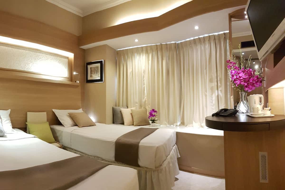 A hotel room with two beds, a bedside table with a phone and a vase of flowers, a large window with curtains, and a mounted TV.