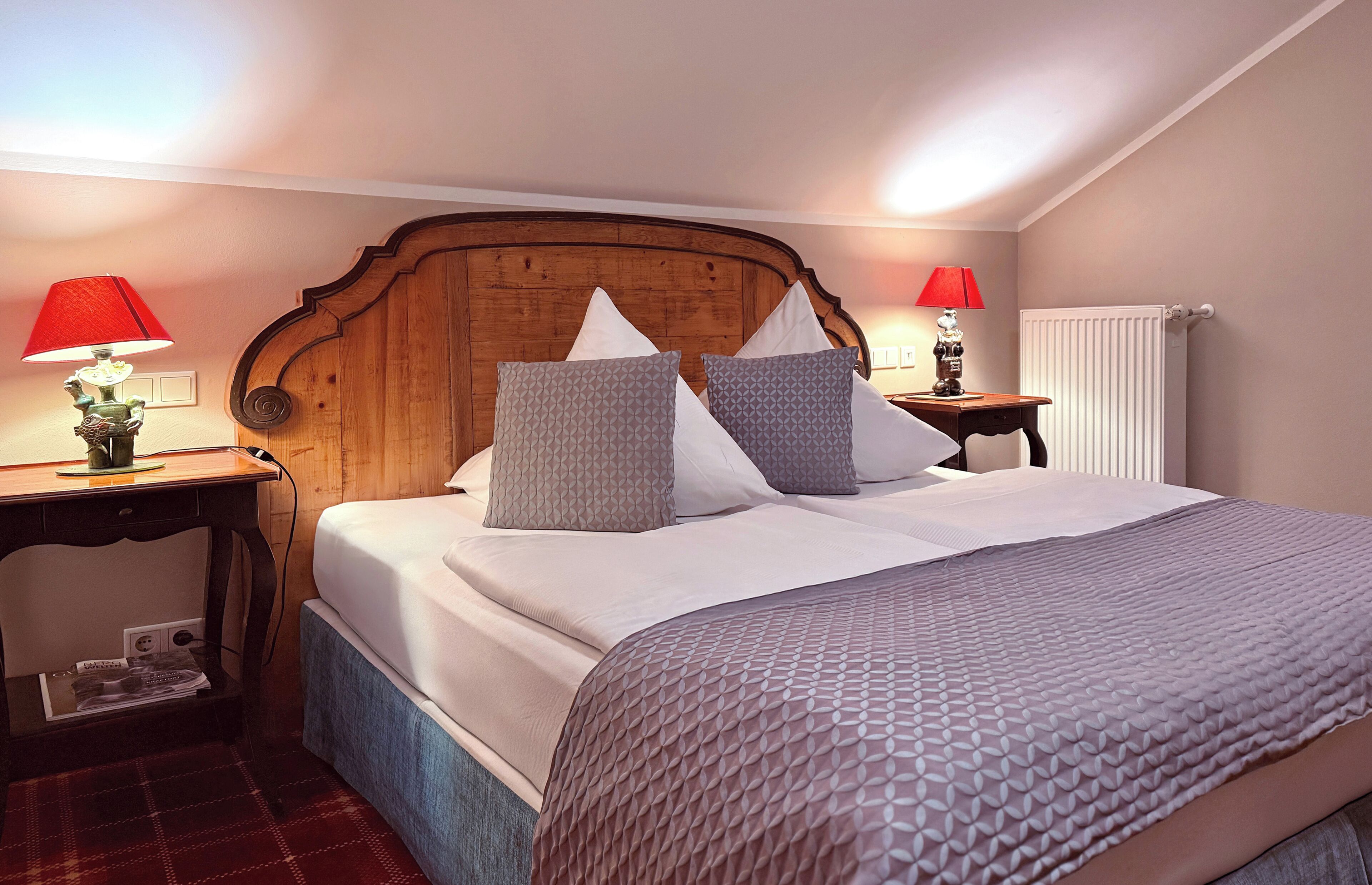 Een net opgemaakt bed met een houten hoofdbord, twee nachtkastjes met lampen en een radiator aan de muur.