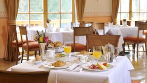 A breakfast table set with croissants, fruit, eggs, and orange juice in a well-lit dining area with large windows. - Romantik Alpenhotel Waxenstein (Grainau)