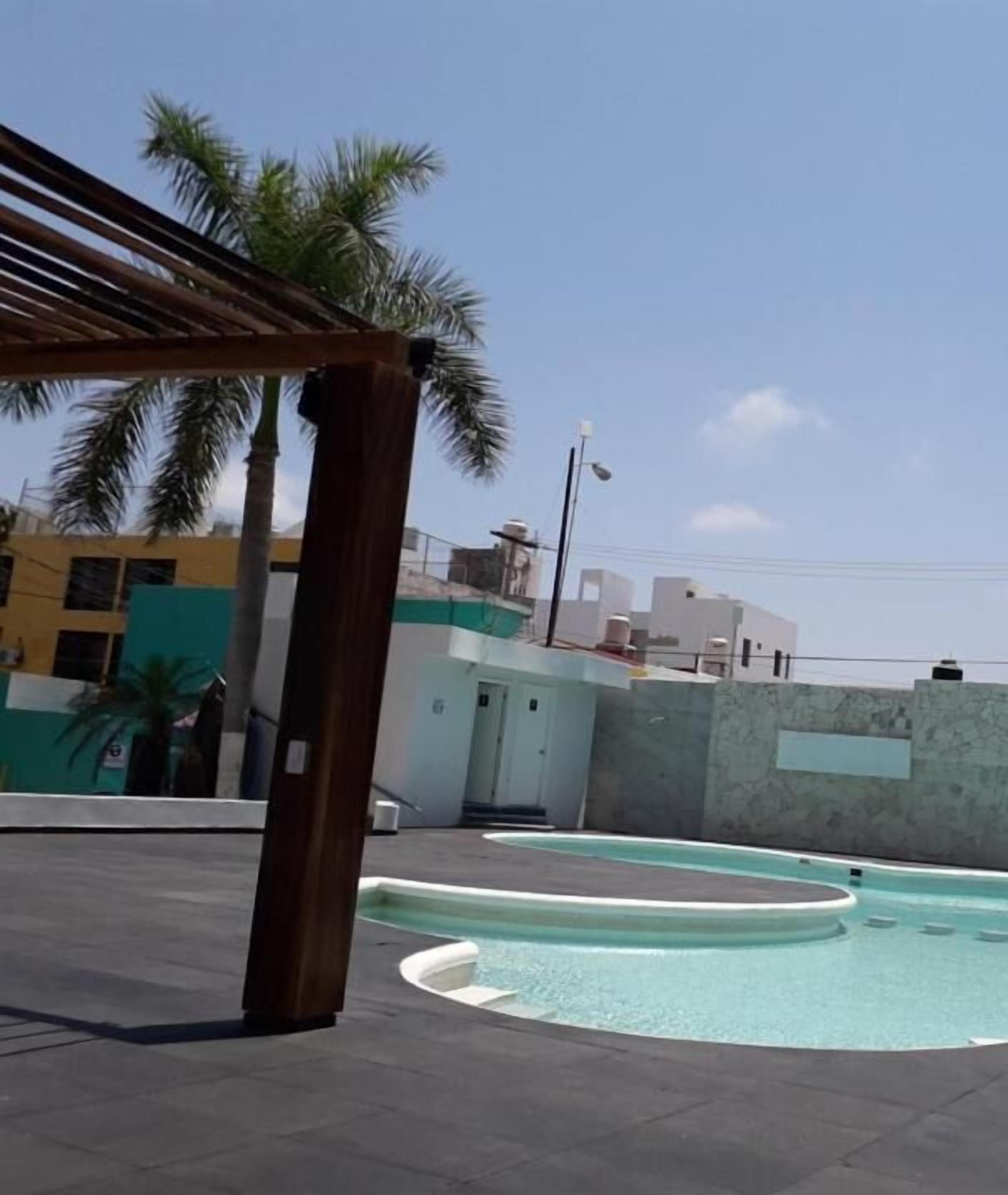 Zona de piscina con una pérgola, palmeras y edificios al fondo.