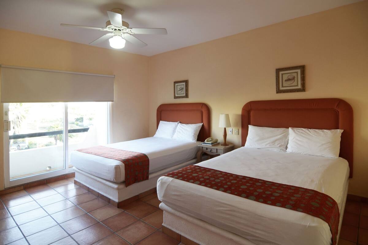 Habitación de hotel con dos camas, ventilador de techo, ventana con vistas y una mesita con lámpara.