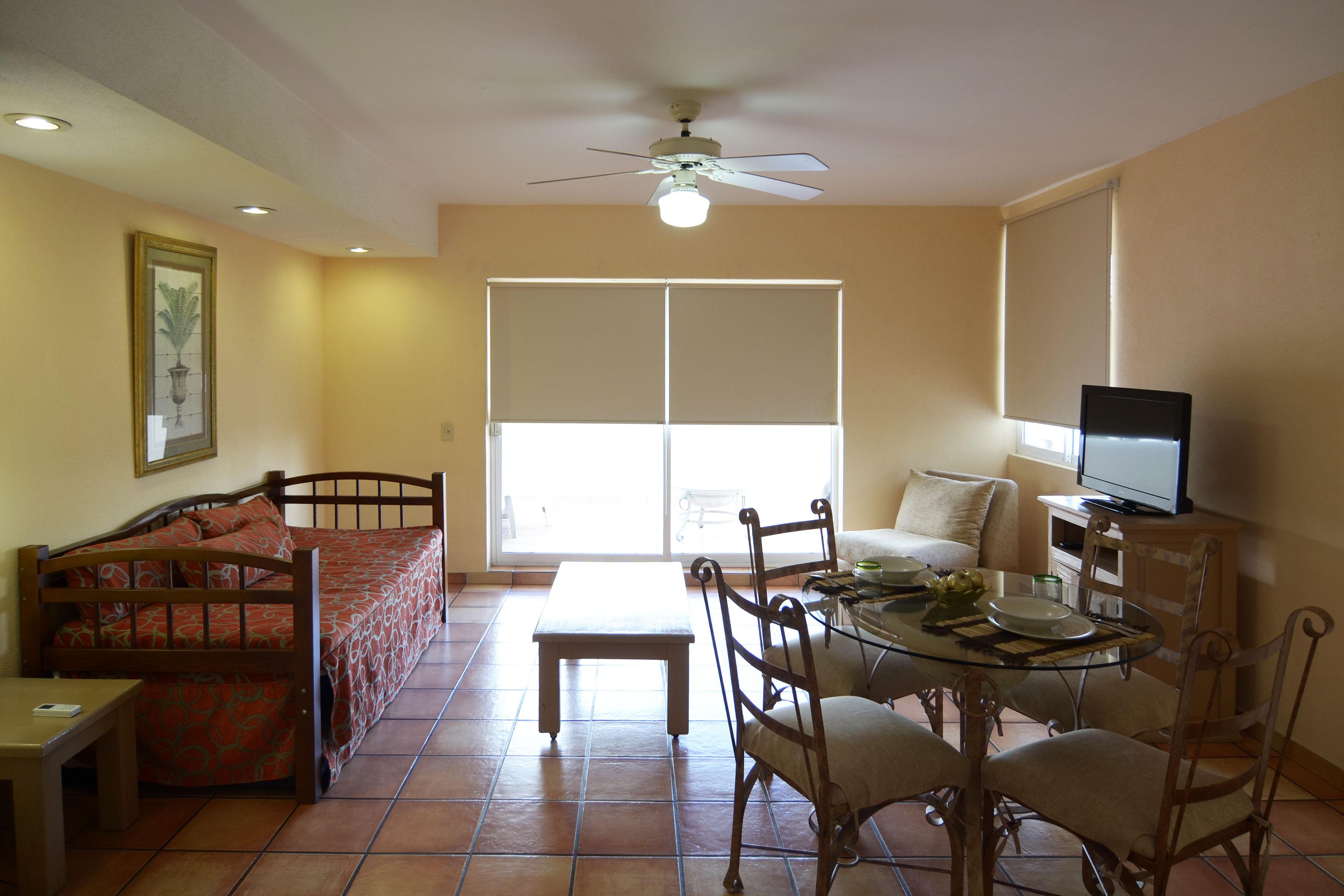 A room with a bed, a dining table with chairs, a ceiling fan, and a television.
